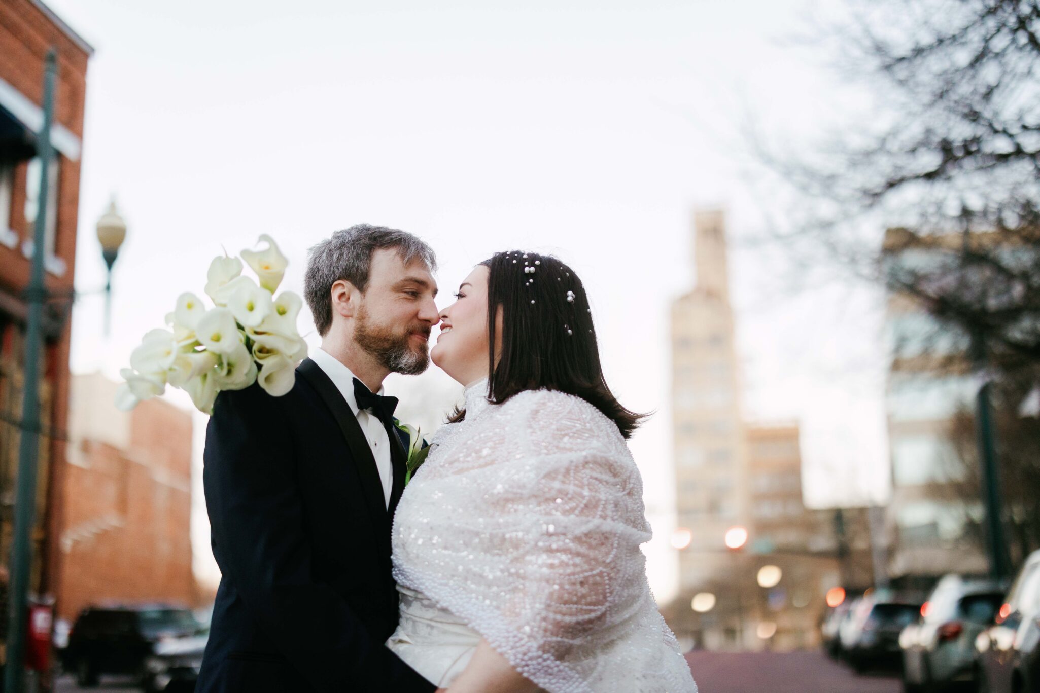 A bride and groom in wedding attire stand close together on a city street, about to kiss. The bride holds white flowers and wears a shawl; snow or confetti rests in her dark hair during their Downtown Asheville wedding.
