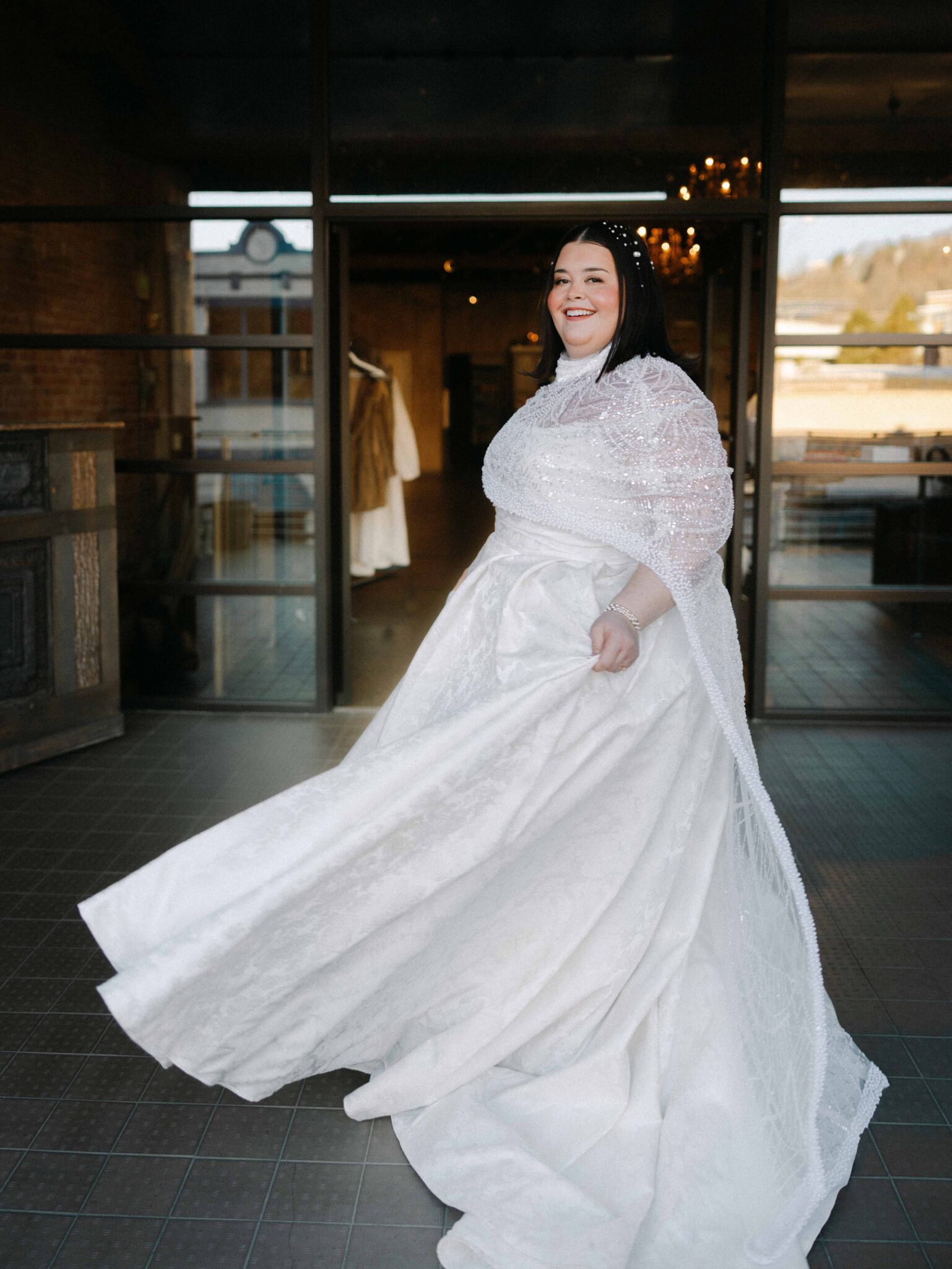 A smiling bride in a white gown twirls indoors at The Venue Downtown Asheville Weddings, her dress flowing gracefully. She has dark hair and wears a pearl headband, with glass doors and warm lighting in the background.