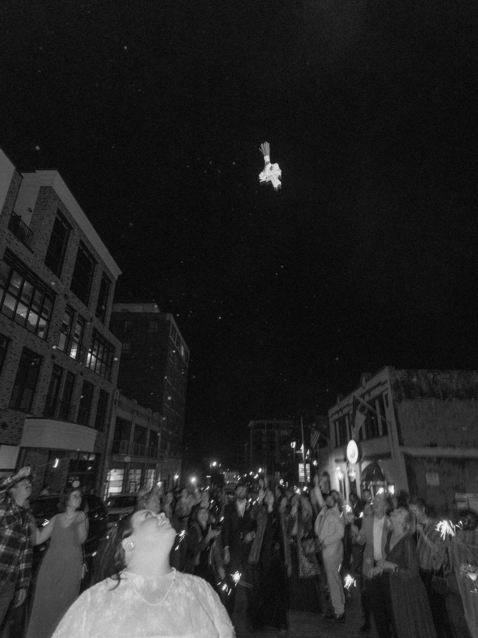 A bride in a wedding dress laughs while looking up as a bouquet flies through the air above a nighttime crowd gathered on a city street.
