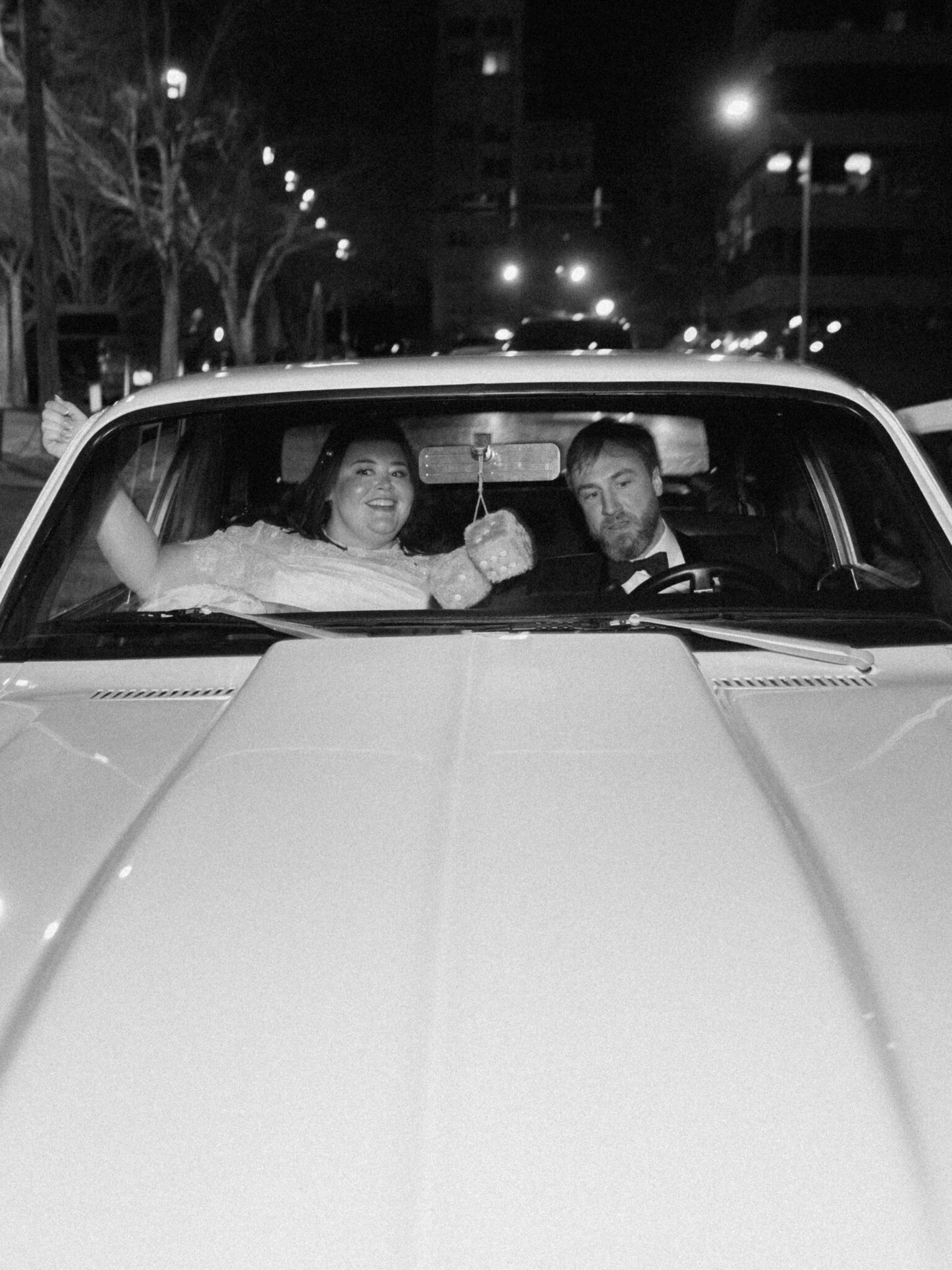A joyful couple sits in the front seat of a classic car at night. The woman, in a white dress, smiles and raises her bouquet, while the man in a suit drives. Streetlights and city buildings are visible in the background.