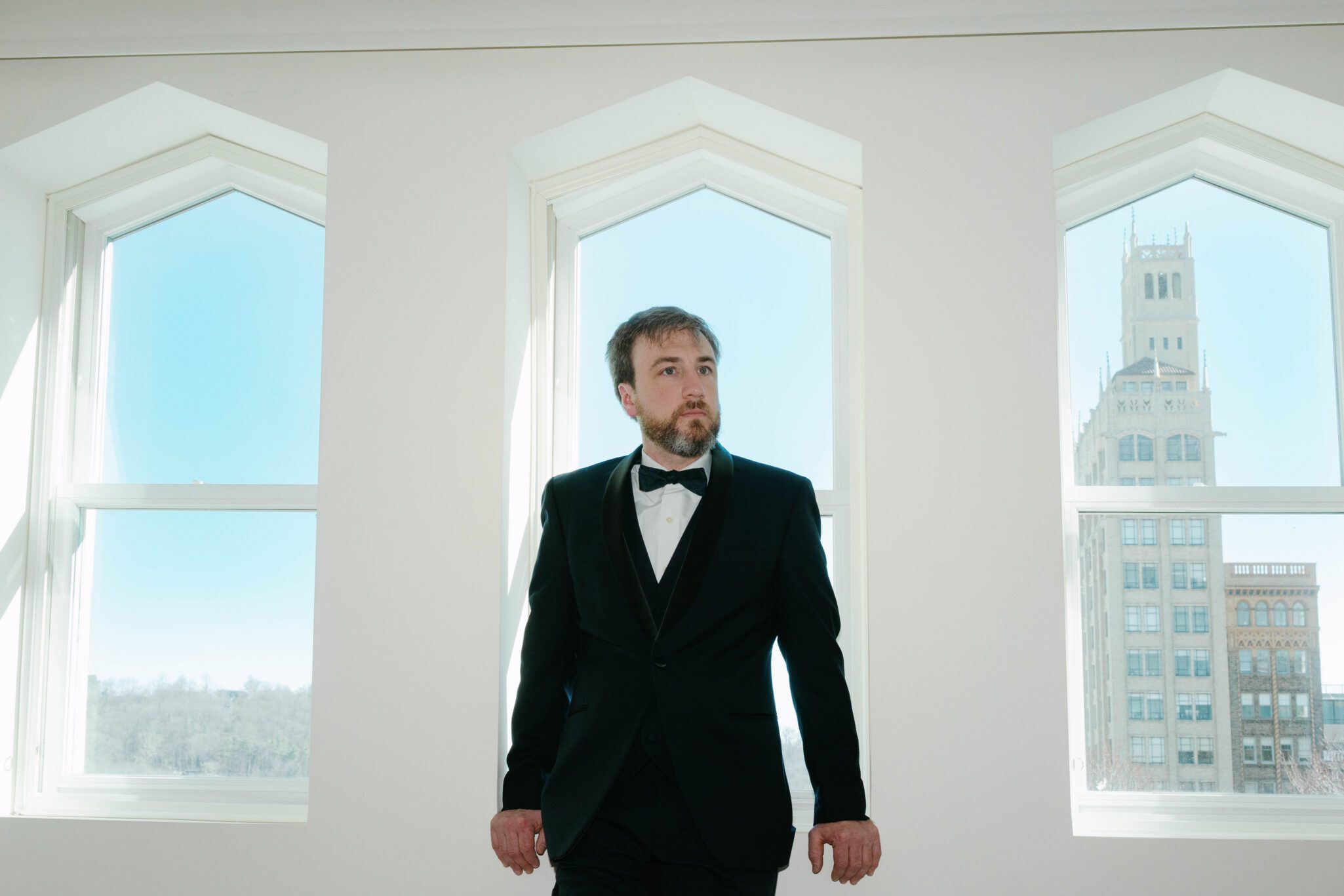 A man in a black tuxedo stands indoors between two tall windows with white frames. Sunlight shines through, and a tall building is visible outside against a clear blue sky.