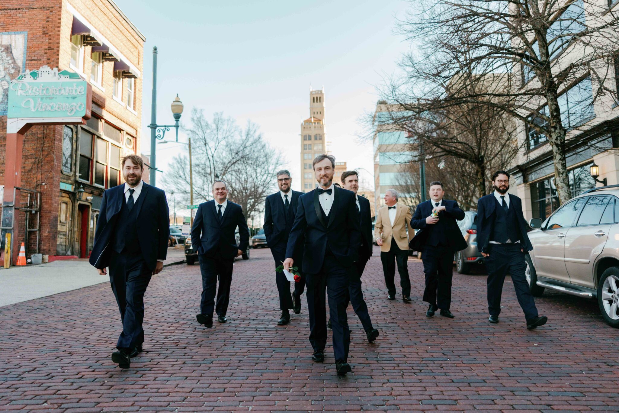 A group of men in formal suits, including one in a tuxedo holding a rose, walk confidently down a brick street in a city with bare trees and historic buildings in the background.