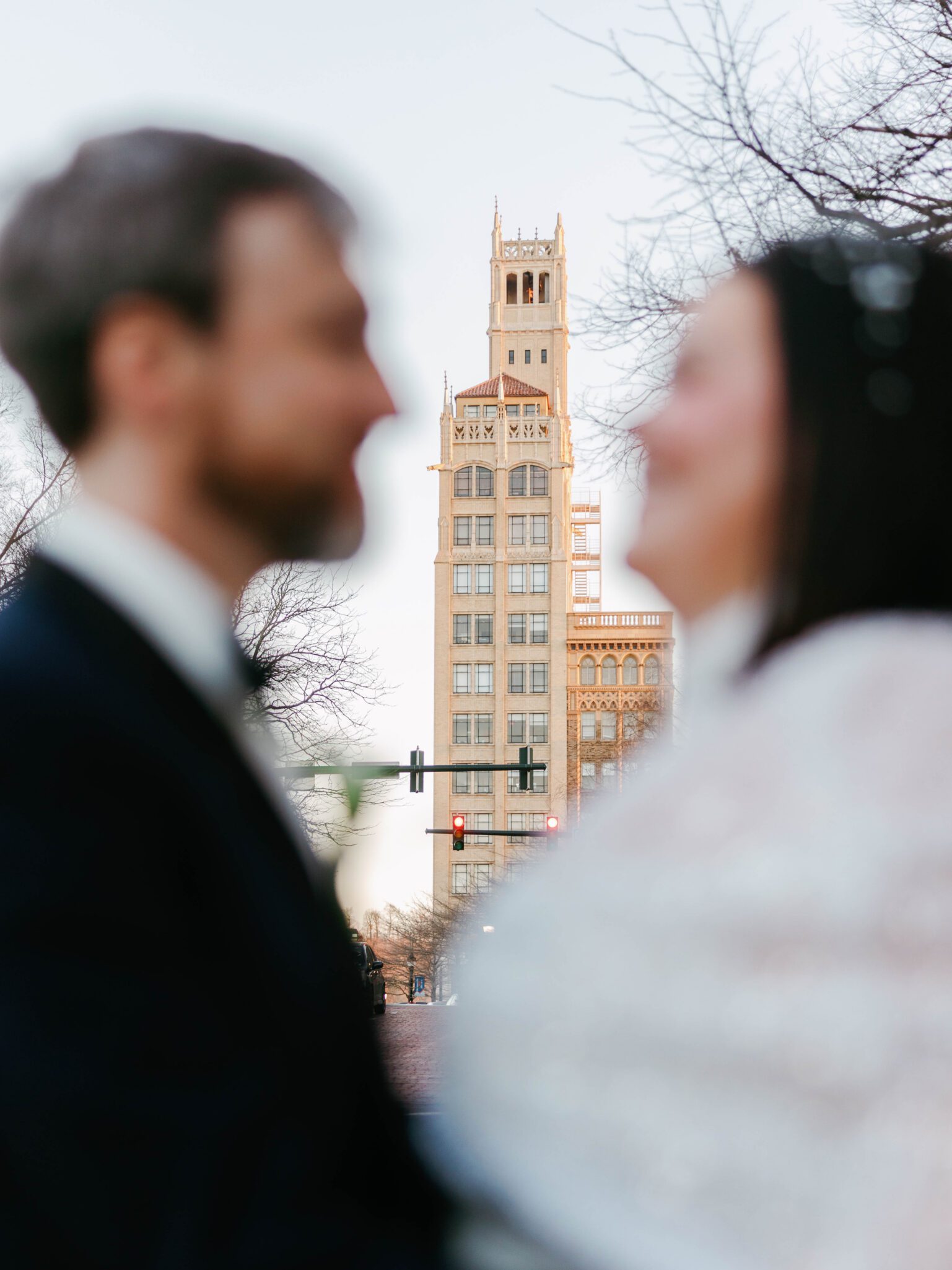 A blurred couple faces each other in the foreground, with a tall, detailed building in sharp focus behind them, framed by bare tree branches and a city street with traffic lights.
