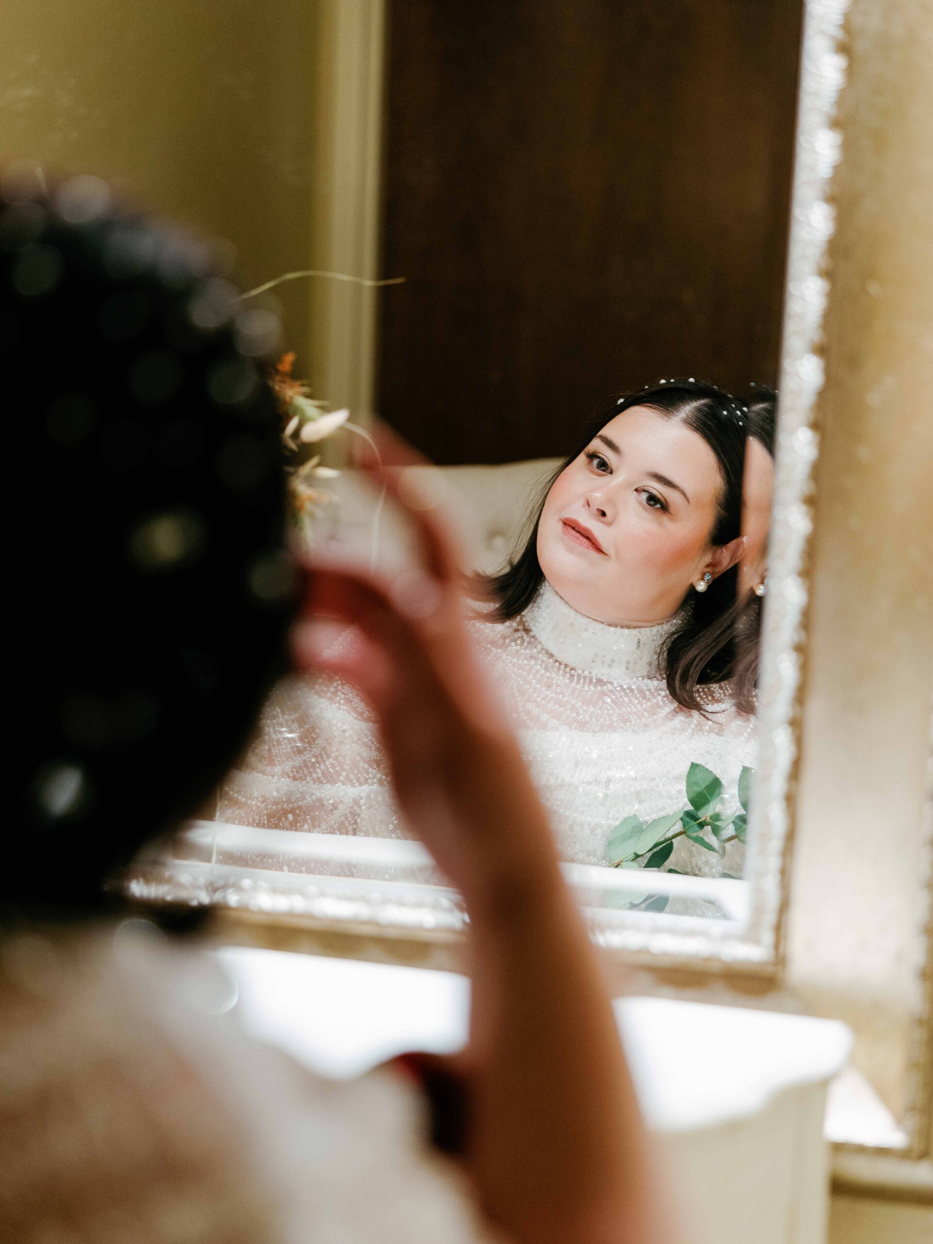 A bride with dark hair in a white turtleneck looks thoughtfully at her reflection in a mirror inside The Venue Downtown Asheville, resting her head on her hand as another person helps her get ready in the foreground.