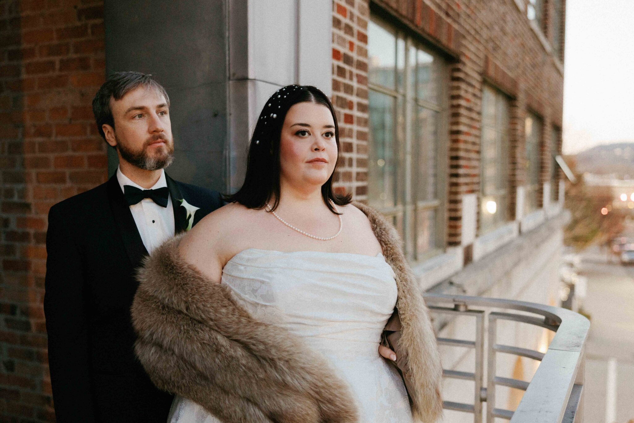 A bride in a white dress and fur stole stands on a balcony beside a groom in a black tuxedo at The Venue Downtown Asheville Weddings. They both look ahead with serious expressions, with brick walls and city buildings in the background.