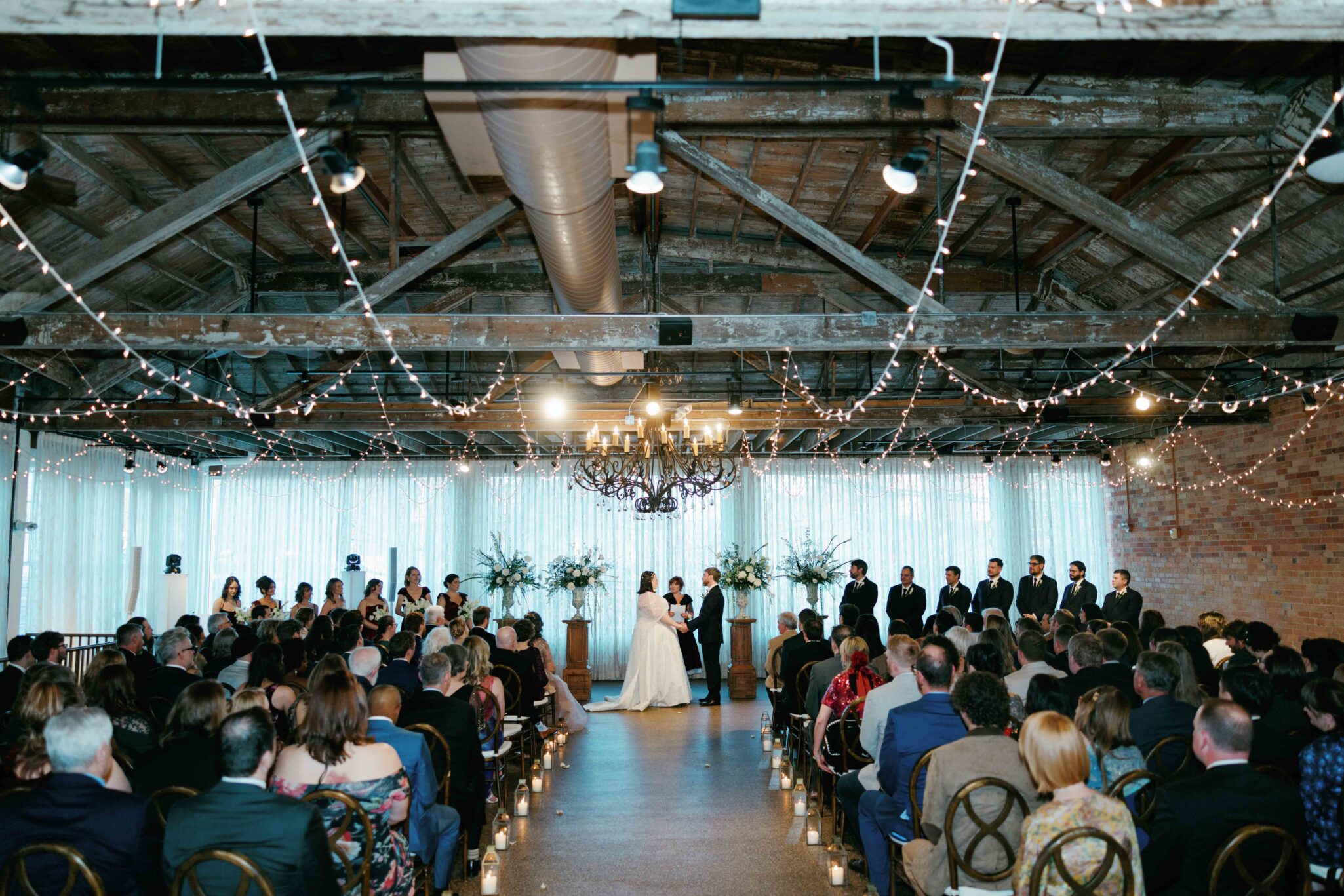A wedding ceremony in a rustic venue with exposed beams and string lights. The bride and groom stand at the altar, surrounded by their wedding party, while guests sit in rows watching the ceremony.