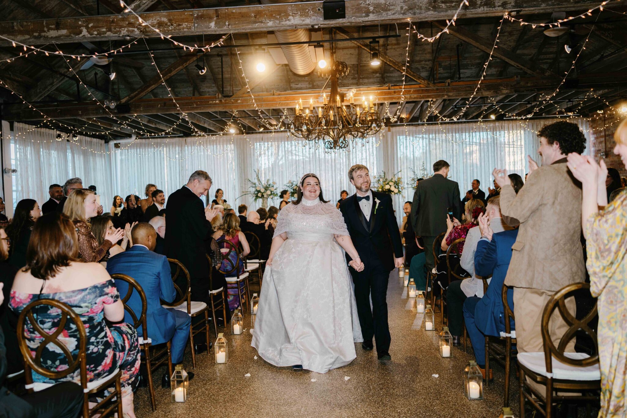 A newlywed couple walks down the aisle hand in hand at an indoor wedding ceremony, surrounded by clapping guests. String lights and candles line the aisle, and a chandelier hangs from the ceiling.