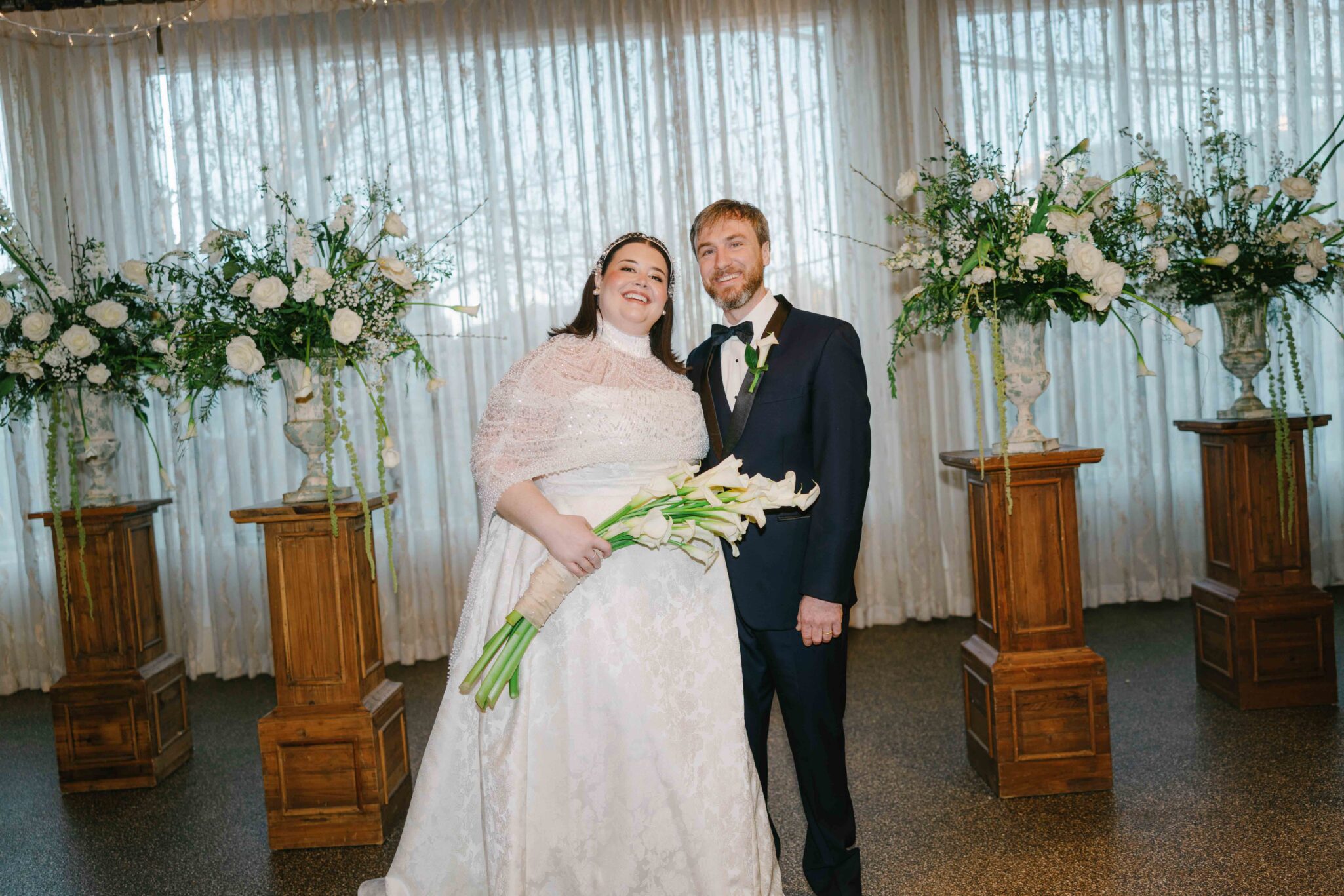 A bride in a white gown holding a bouquet stands beside a groom in a black suit, both smiling. The Venue wedding ceremony set up features large floral arrangements and elegant white drapery in the background.