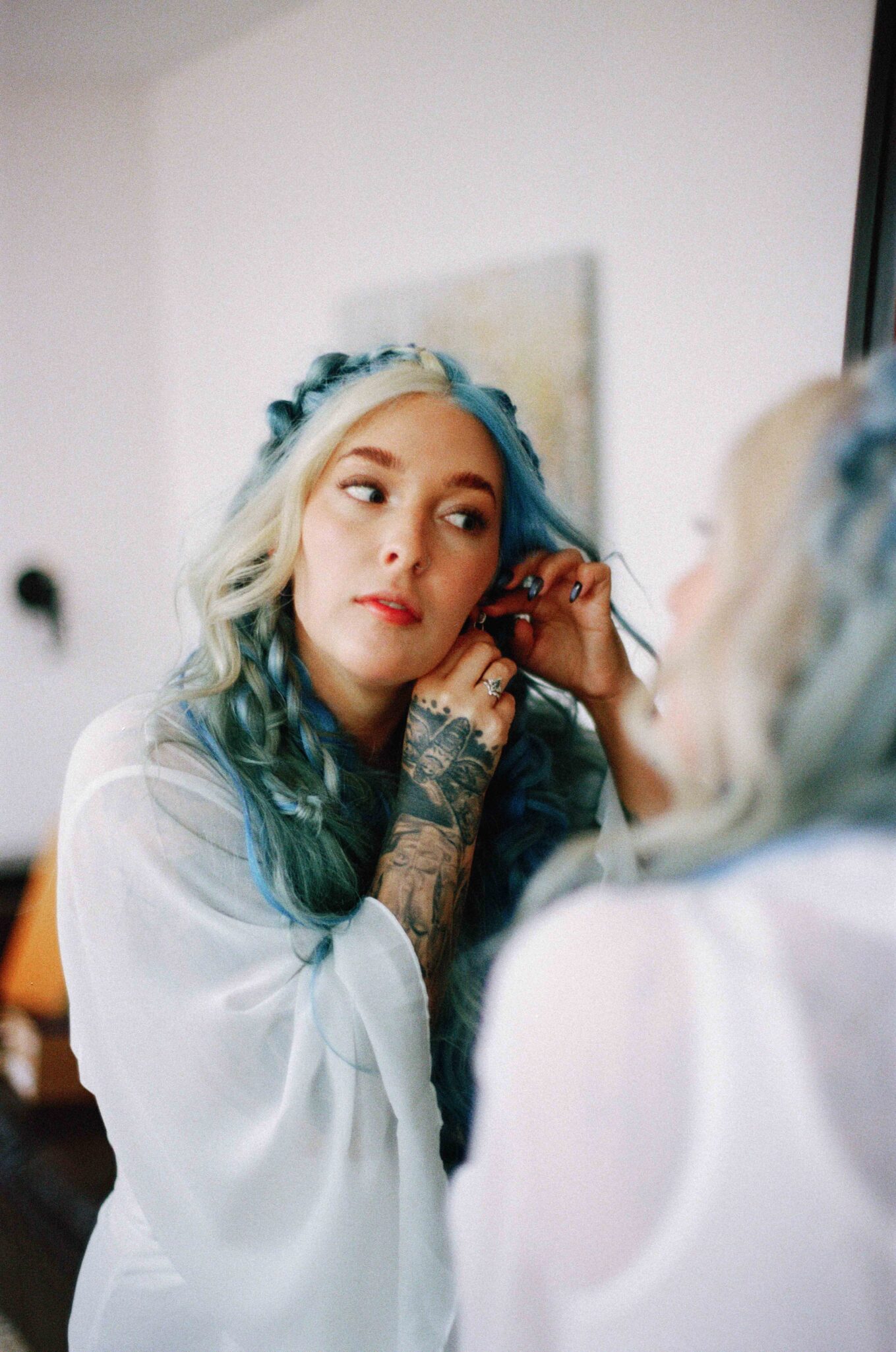 asheville wedding photographer A woman with long blue and blonde hair and tattoos adjusts her earring in a white robe, her reflection captured in softly blurred film photography—perfect for the timeless elegance of Biltmore Estate weddings.