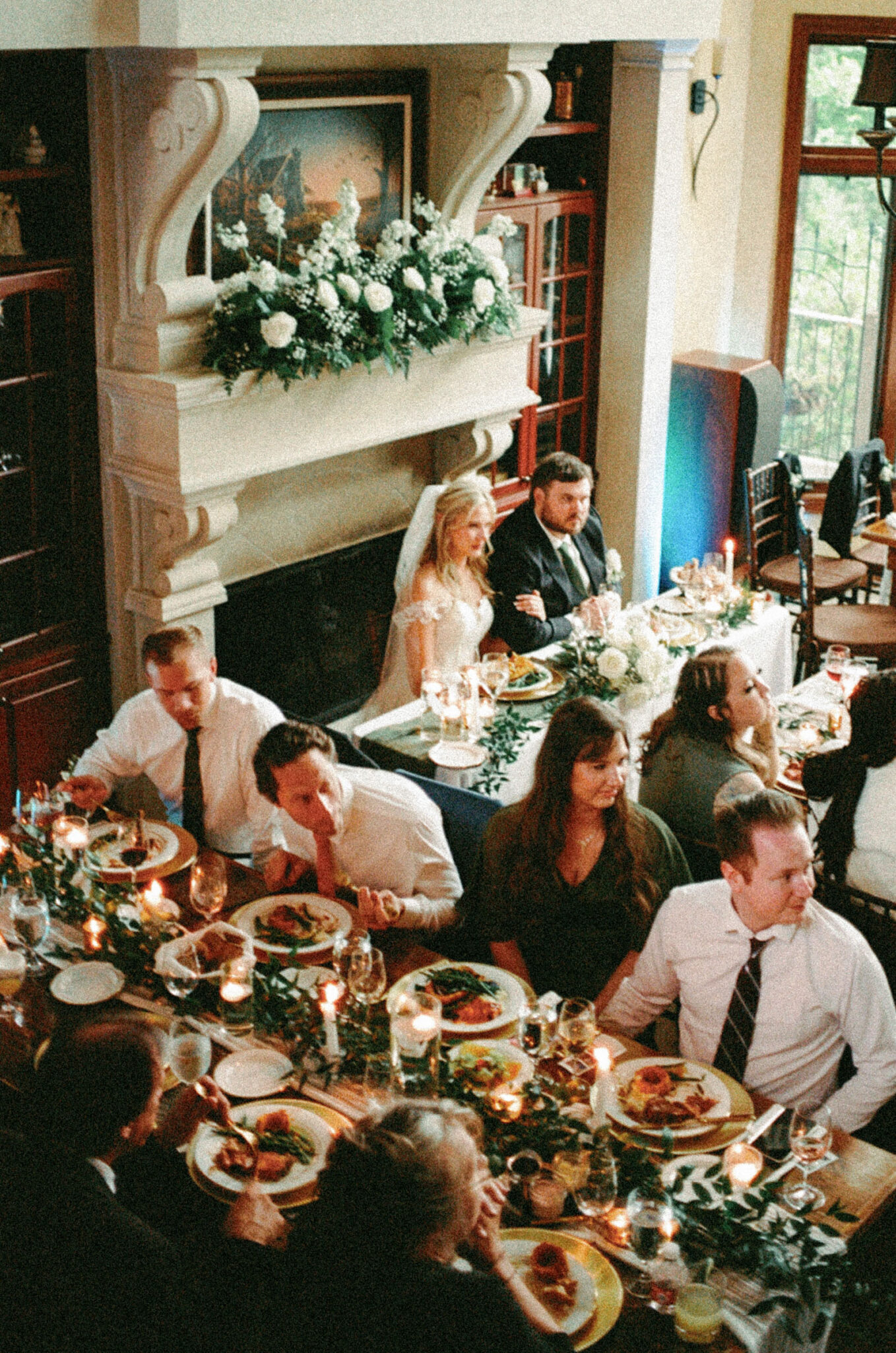 asheville wedding photographer A bride and groom sit at a decorated table near a fireplace, smiling during their wedding reception at the Biltmore Estate. Guests dine below, with candlelight and floral arrangements captured beautifully in film photography.