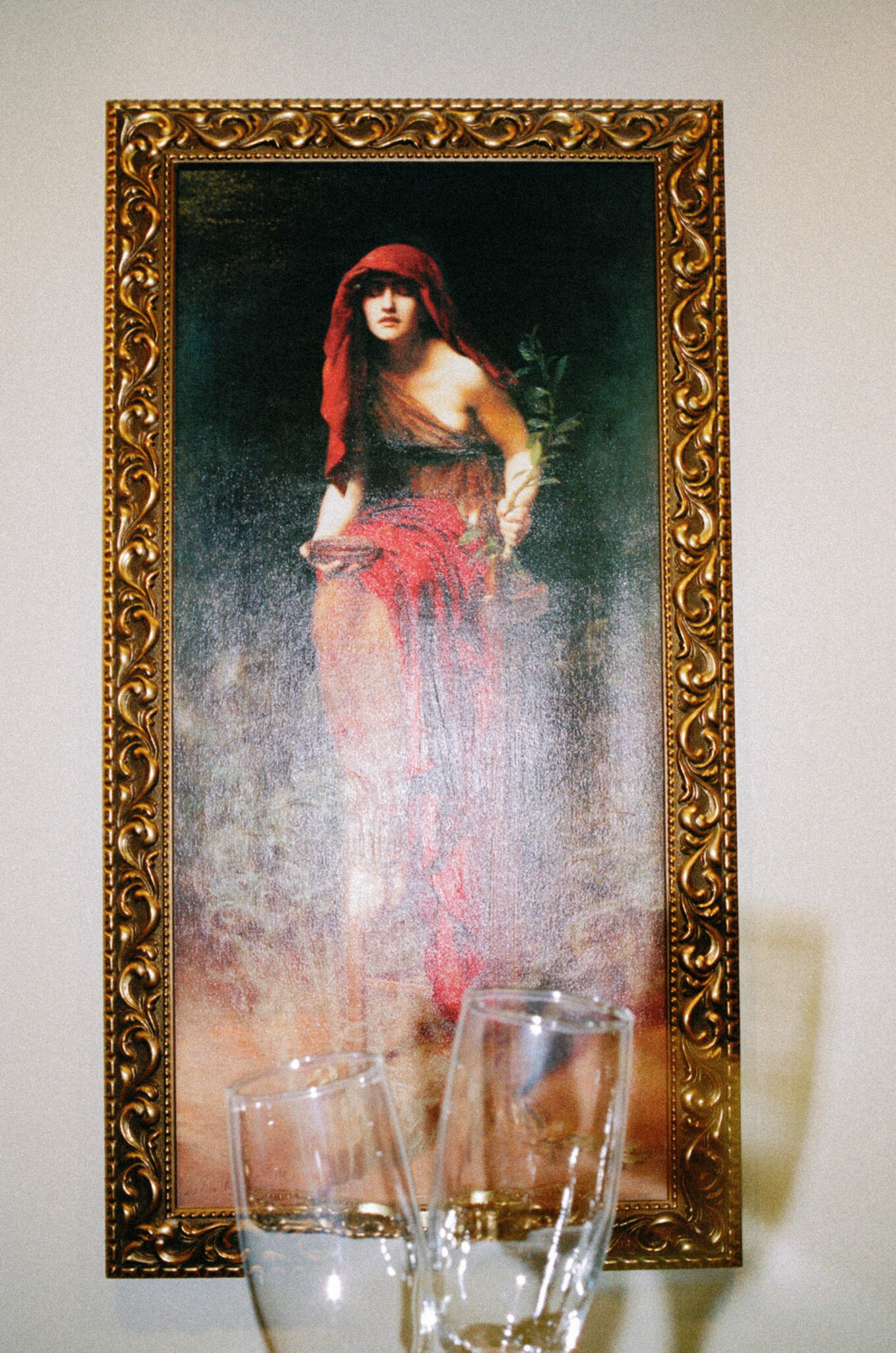 asheville wedding photographer A framed painting of a woman in a red headscarf and dress holding greenery, with two empty champagne glasses in the foreground, set against a plain wall—captured with the timeless elegance of film photography.