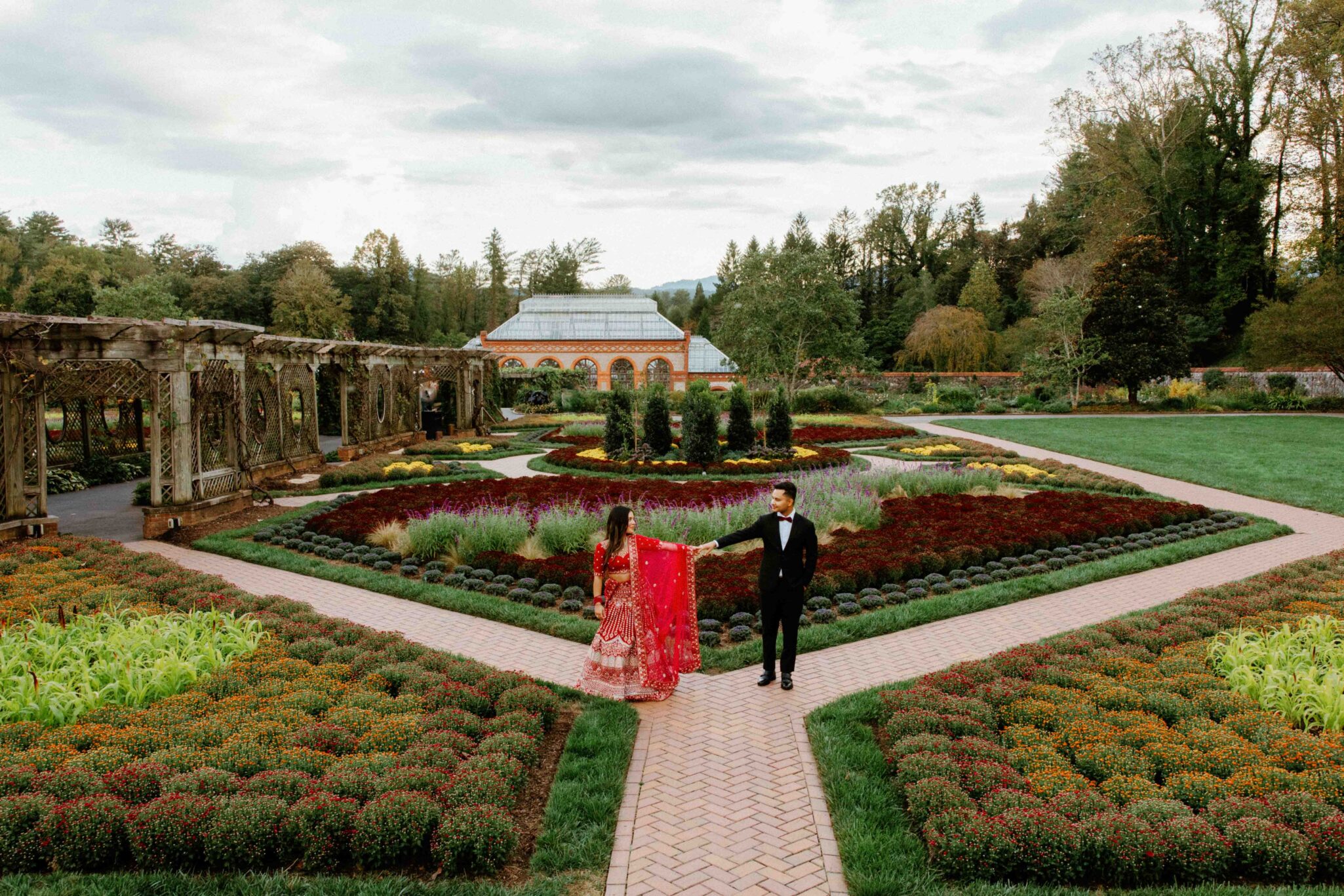 A couple in formal attire, with the woman in a red dress and the man in a black suit, walk hand in hand along a path at The Biltmore Estate’s beautifully landscaped garden, featuring colorful geometric flower beds and a greenhouse in the background.