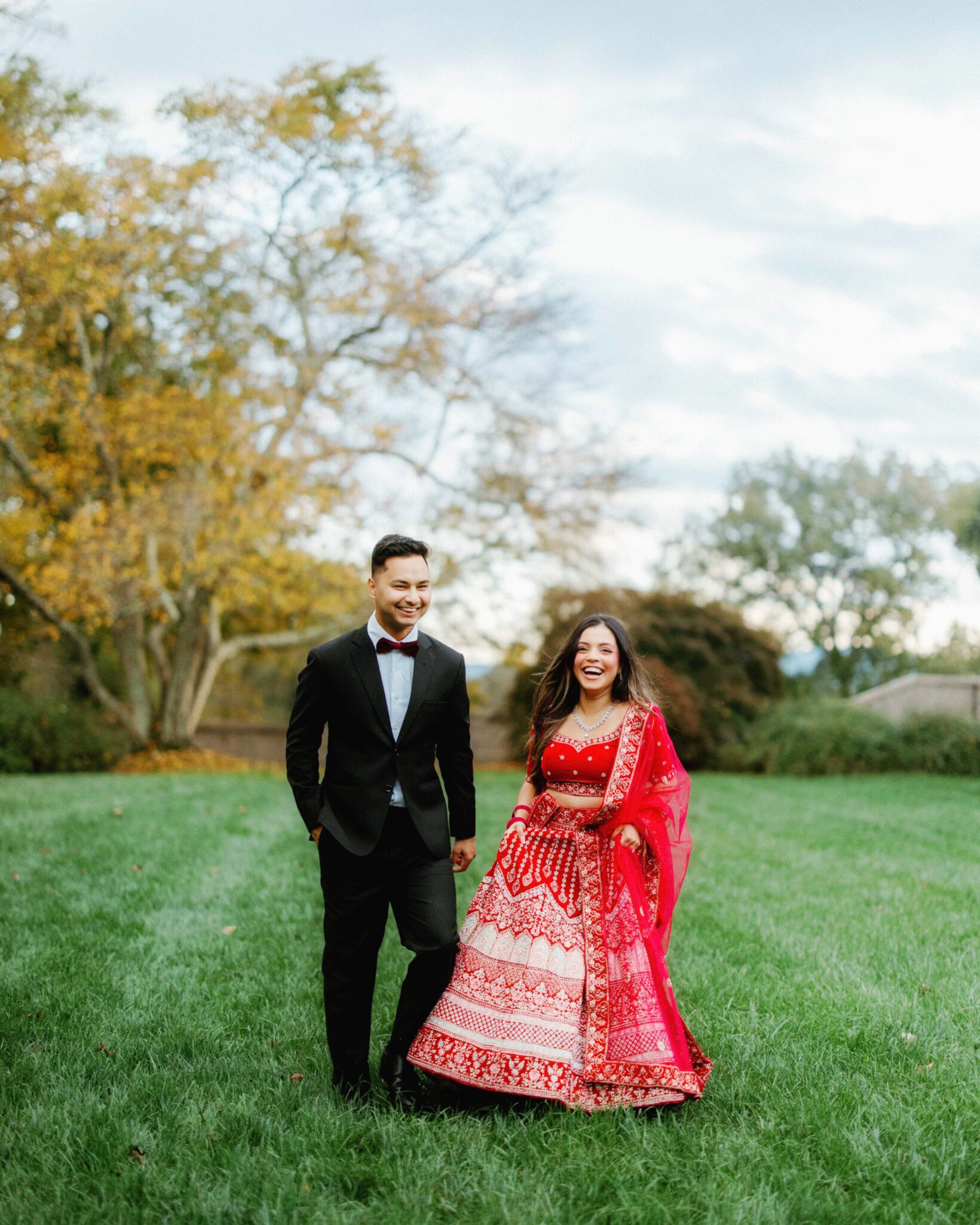 A man in a black suit and a woman in a red and gold traditional dress walk together on green grass, smiling, with trees and a cloudy sky in the background.