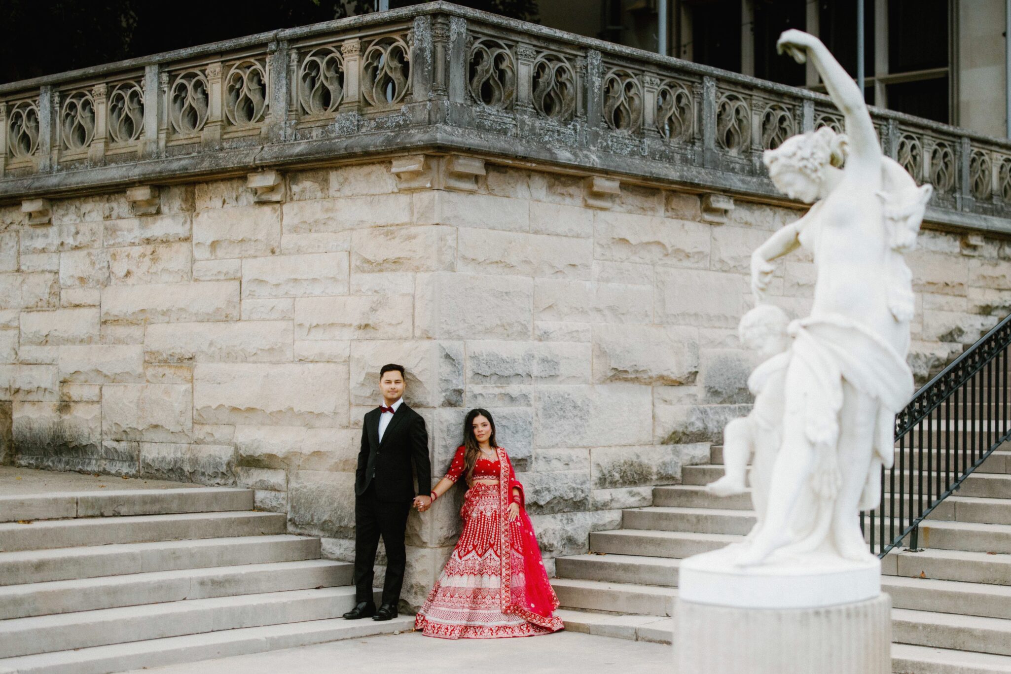 A couple stands on stone steps holding hands; the man wears a black suit and bow tie, and the woman wears a red and white traditional dress. A white statue is visible in the foreground.