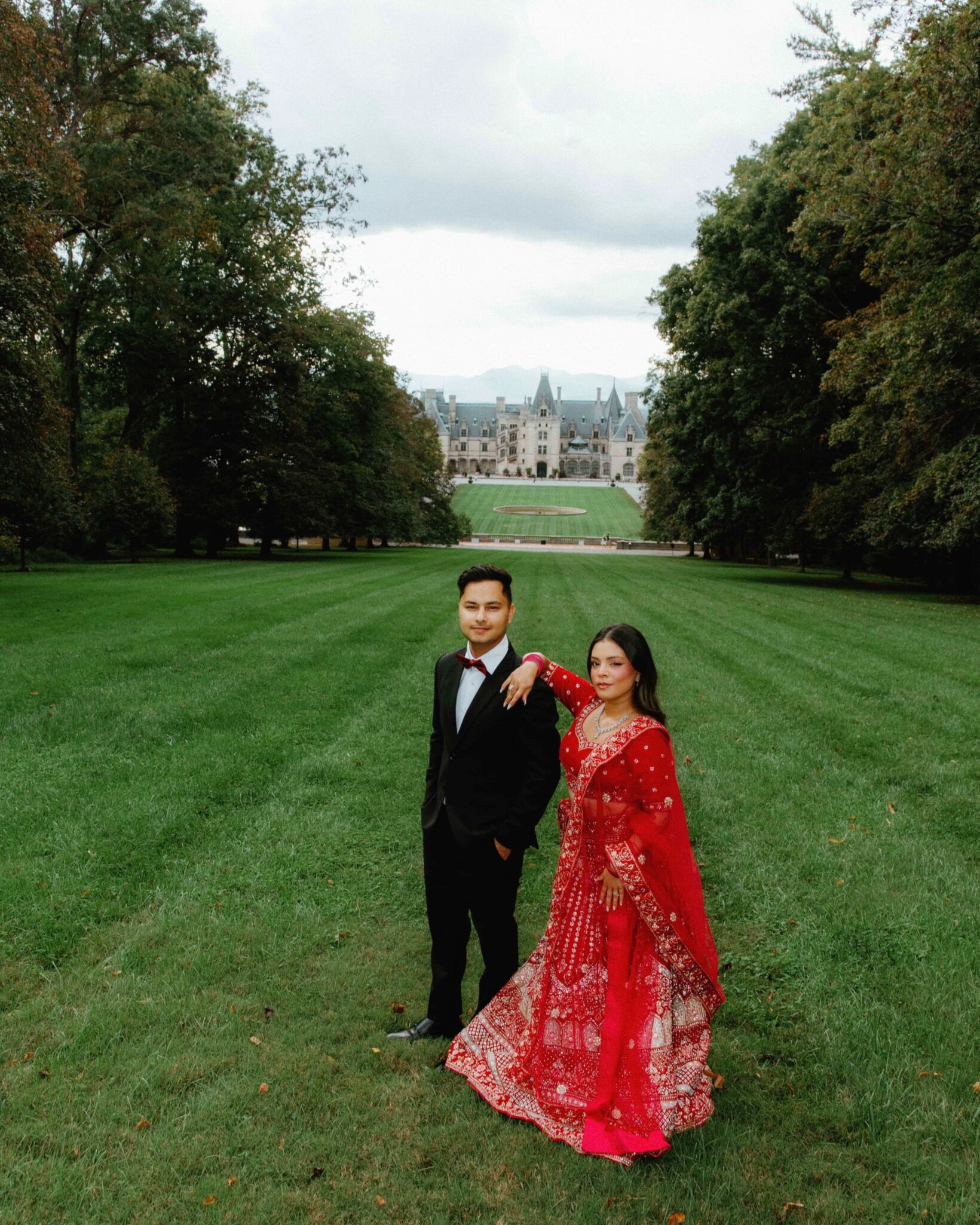 A man in a black suit and a woman in a red traditional outfit pose together on a large green lawn with The Biltmore Estate and trees providing a grand backdrop.