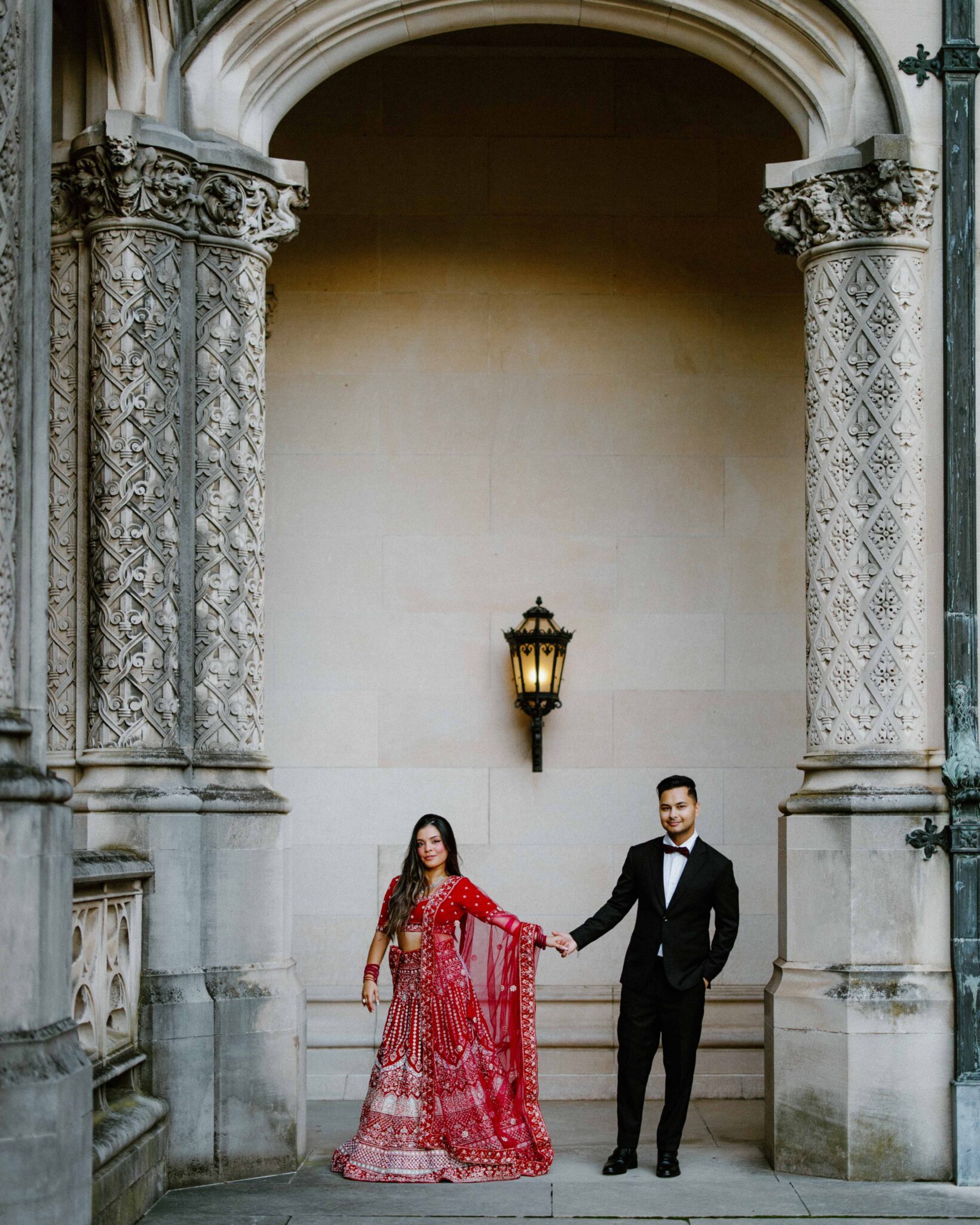 A woman in a red traditional Indian outfit and a man in a black suit hold hands while standing under an ornate stone archway with detailed columns and a hanging lantern.