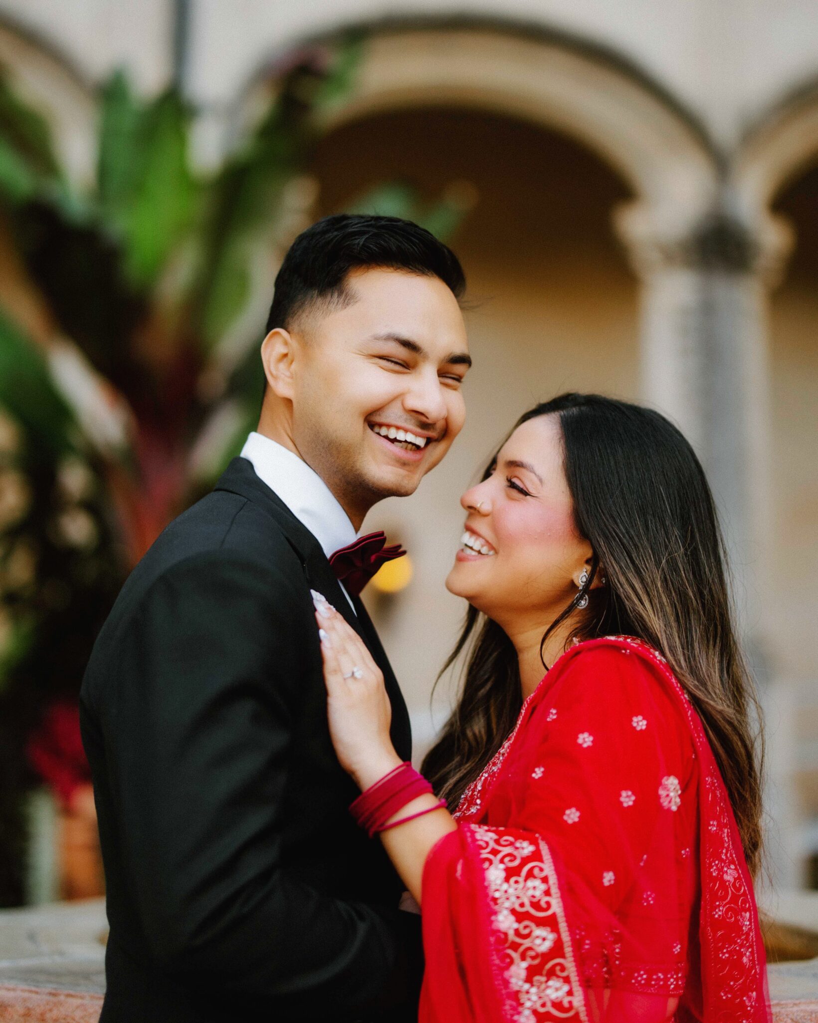 A smiling couple stands close together outdoors. The man wears a black suit with a bow tie, and the woman wears a red traditional outfit with matching bangles. They look happy, with an arched building and greenery in the background.