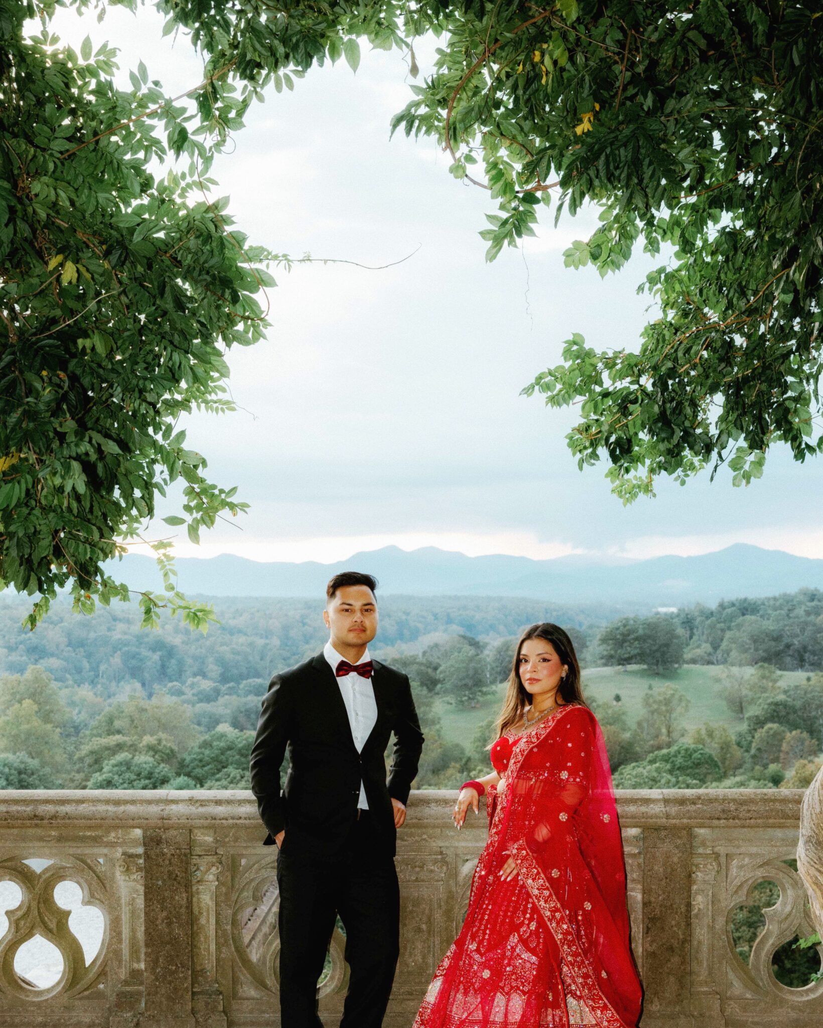 A man in a black suit and red bow tie stands next to a woman in a red sari on a stone balcony, with green trees framing them and mountains visible in the background.