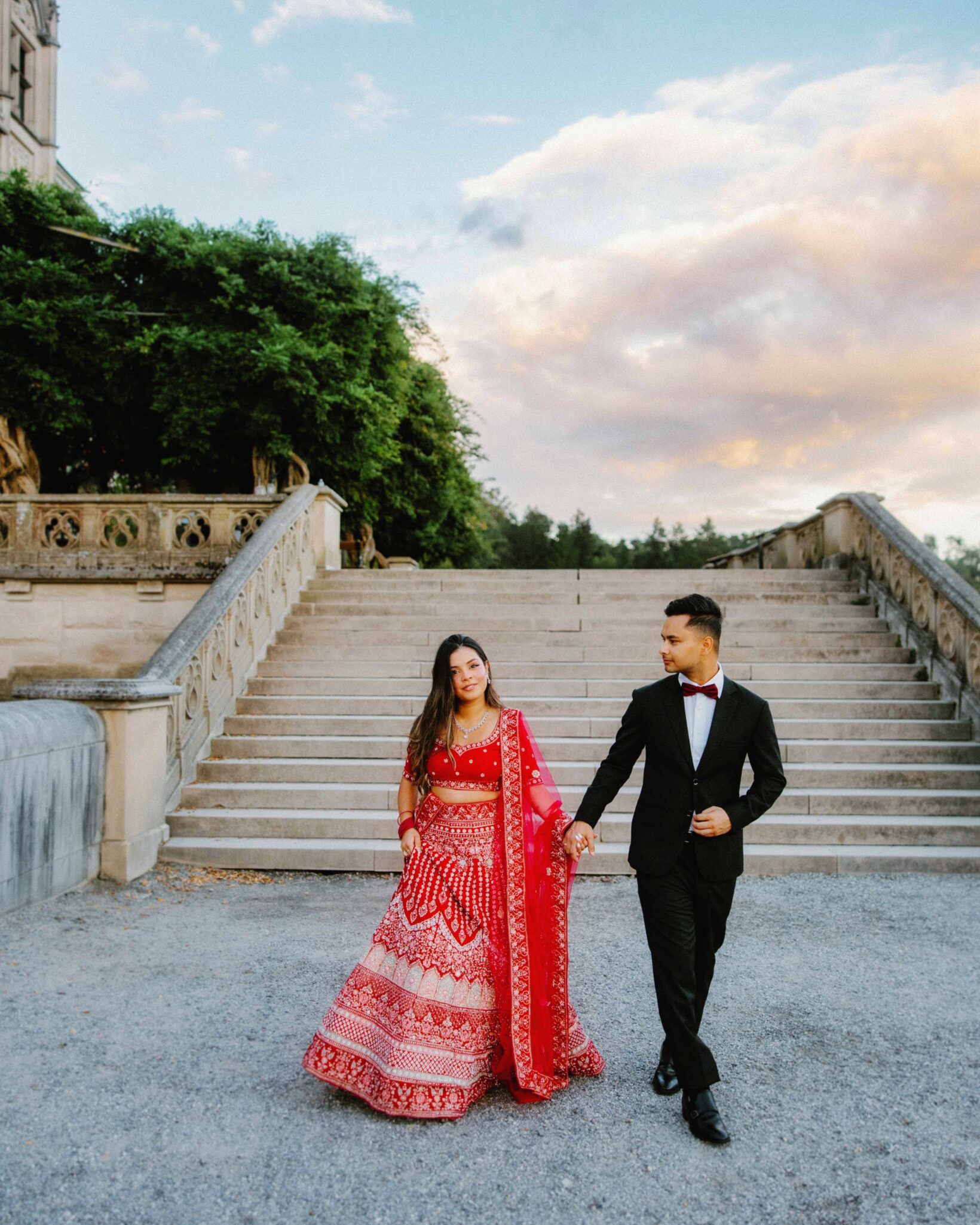 A woman in a red and white lehenga and a man in a black suit and bow tie hold hands, walking in front of grand stone stairs outdoors, with greenery and a partly cloudy sky in the background.
