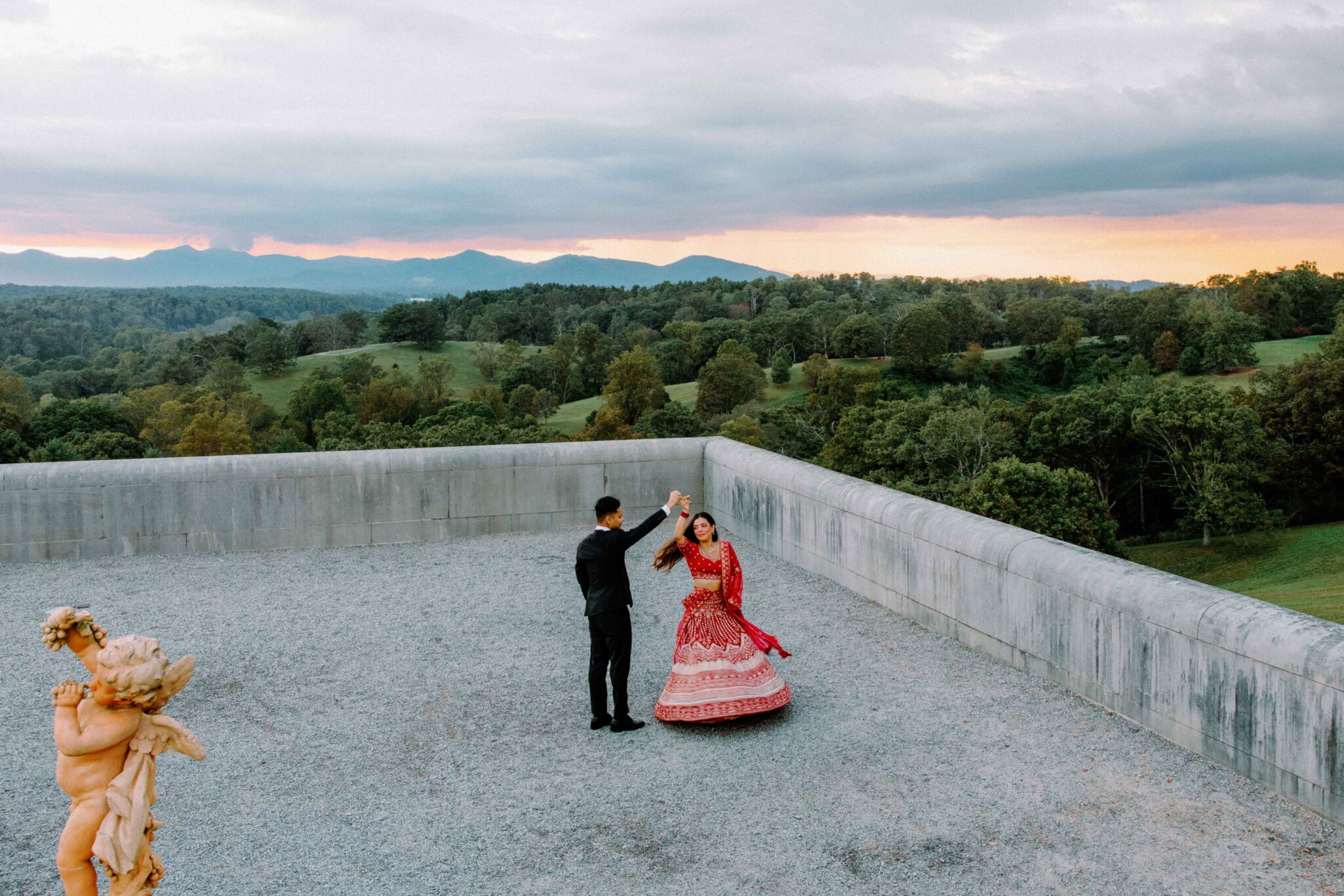 A couple dances on a gravel rooftop near a low concrete wall, surrounded by lush green hills and distant mountains at sunset. The woman wears a red traditional dress; a small statue stands in the foreground.