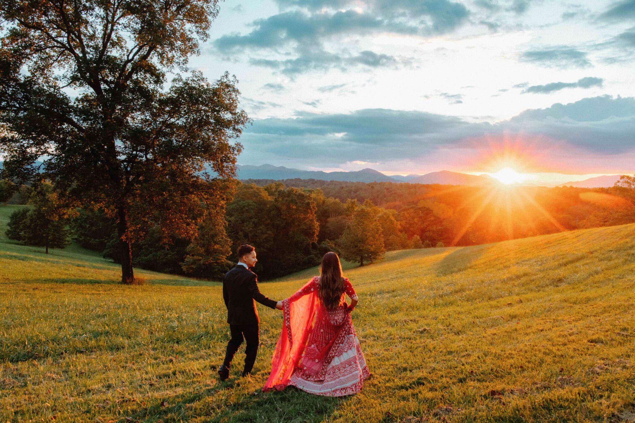 A couple holding hands walks through a sunlit grassy field at sunset. The woman wears a flowing red and white traditional dress, and the man is in a dark suit. Trees and mountains are visible in the background under a partly cloudy sky.