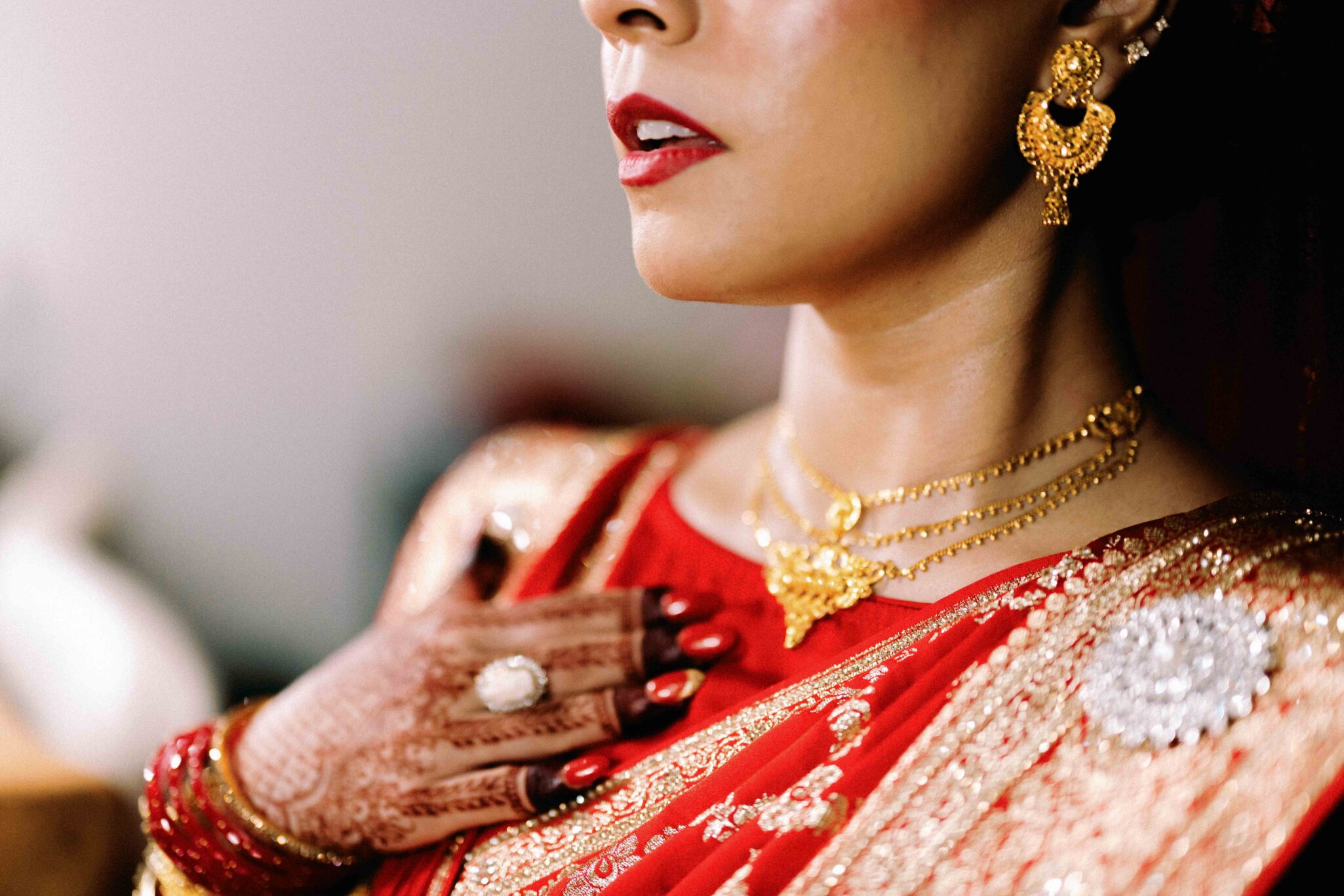 A woman dressed in a red saree with gold embroidery, wearing gold jewelry, including earrings and necklaces. Her hand, decorated with henna and a ring, rests on her chest. Only her lower face and torso are visible.