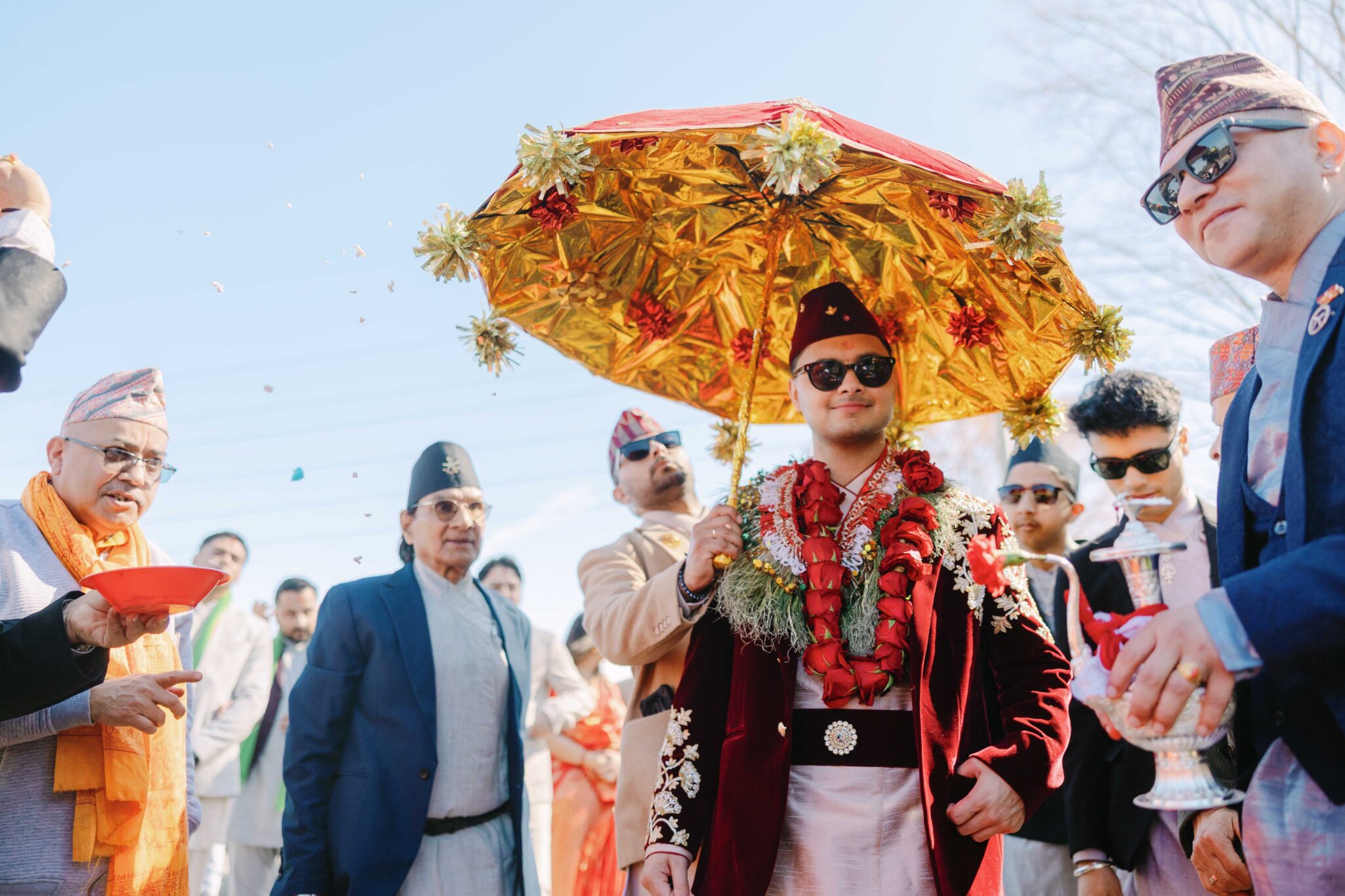 A groom in traditional Nepali attire stands under a decorated golden umbrella, surrounded by well-dressed men, flower petals in the air, during an outdoor wedding ceremony.