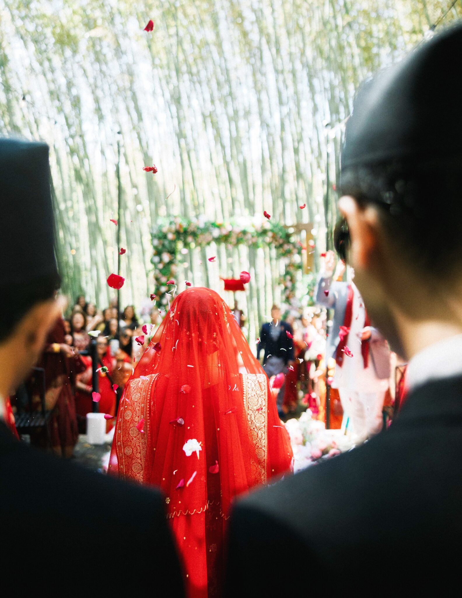 A bride in a red veil walks down an outdoor aisle covered in flower petals, surrounded by greenery and guests, with two men in dark suits visible in the foreground.