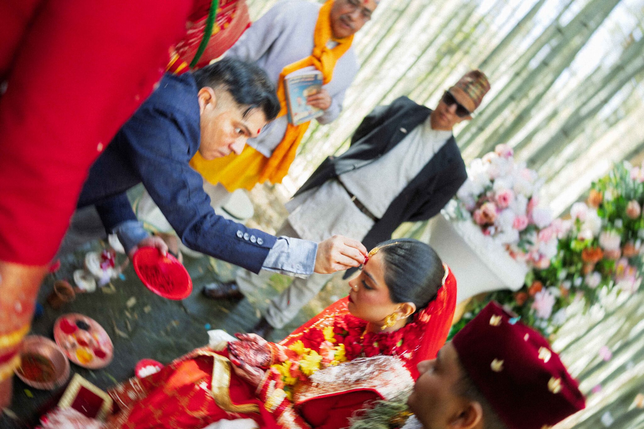 A man applies a red powder to a bride’s forehead during an outdoor wedding ceremony, surrounded by traditionally dressed people, colorful flowers, and trees in the background.