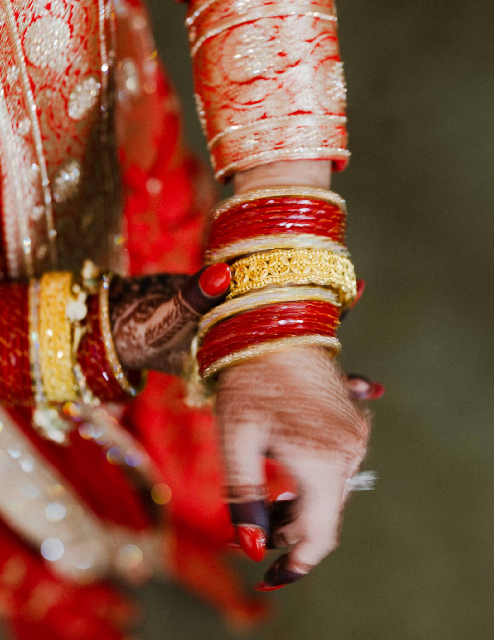 A close-up of a womans hand adorned with red and gold bangles, intricate henna designs, and traditional Indian attire in red and gold fabric.