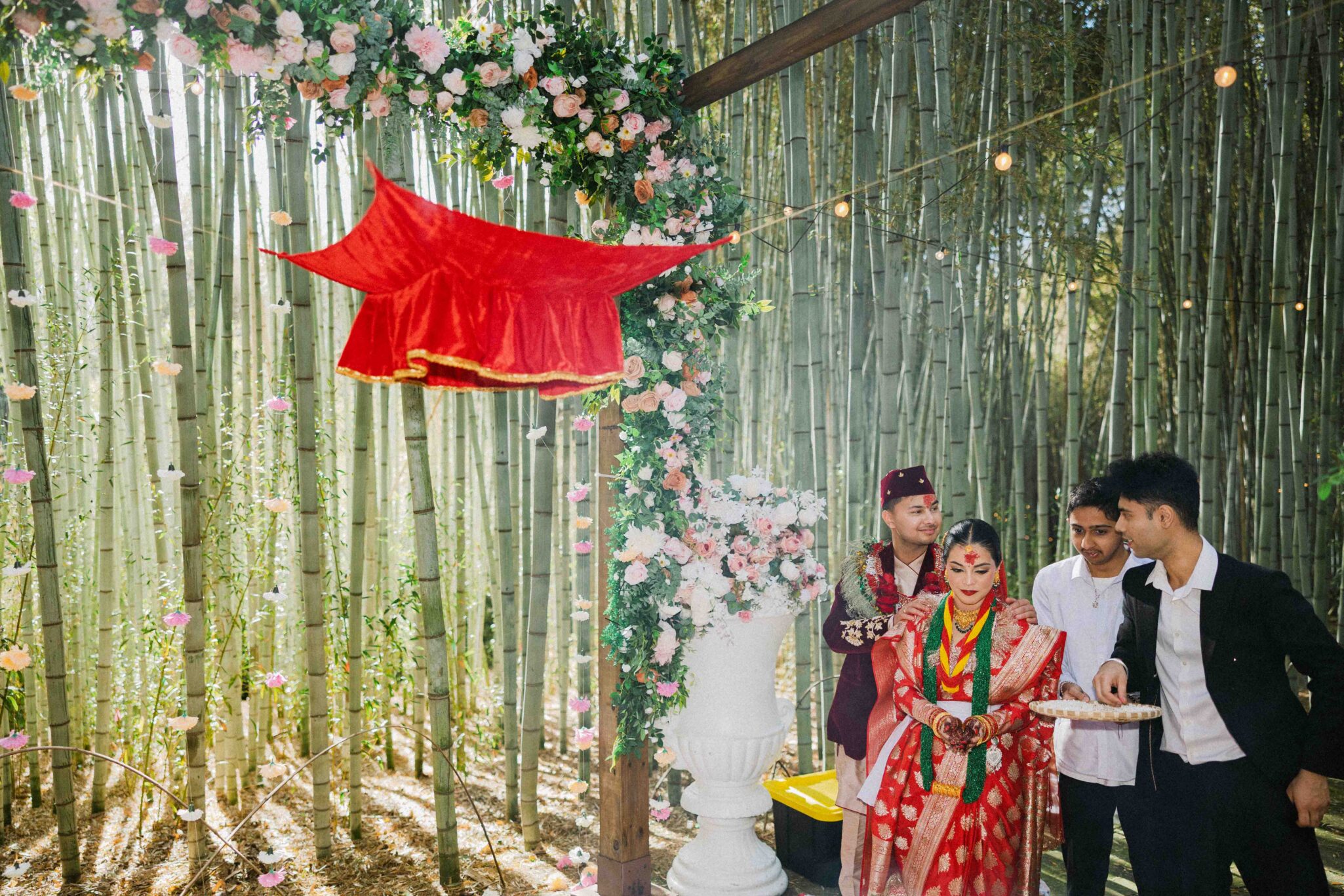 A bride in a red saree and two men stand under a floral arch with a red cloth canopy, set against a bamboo forest. The scene is decorated with flowers and string lights, suggesting an outdoor wedding ceremony.