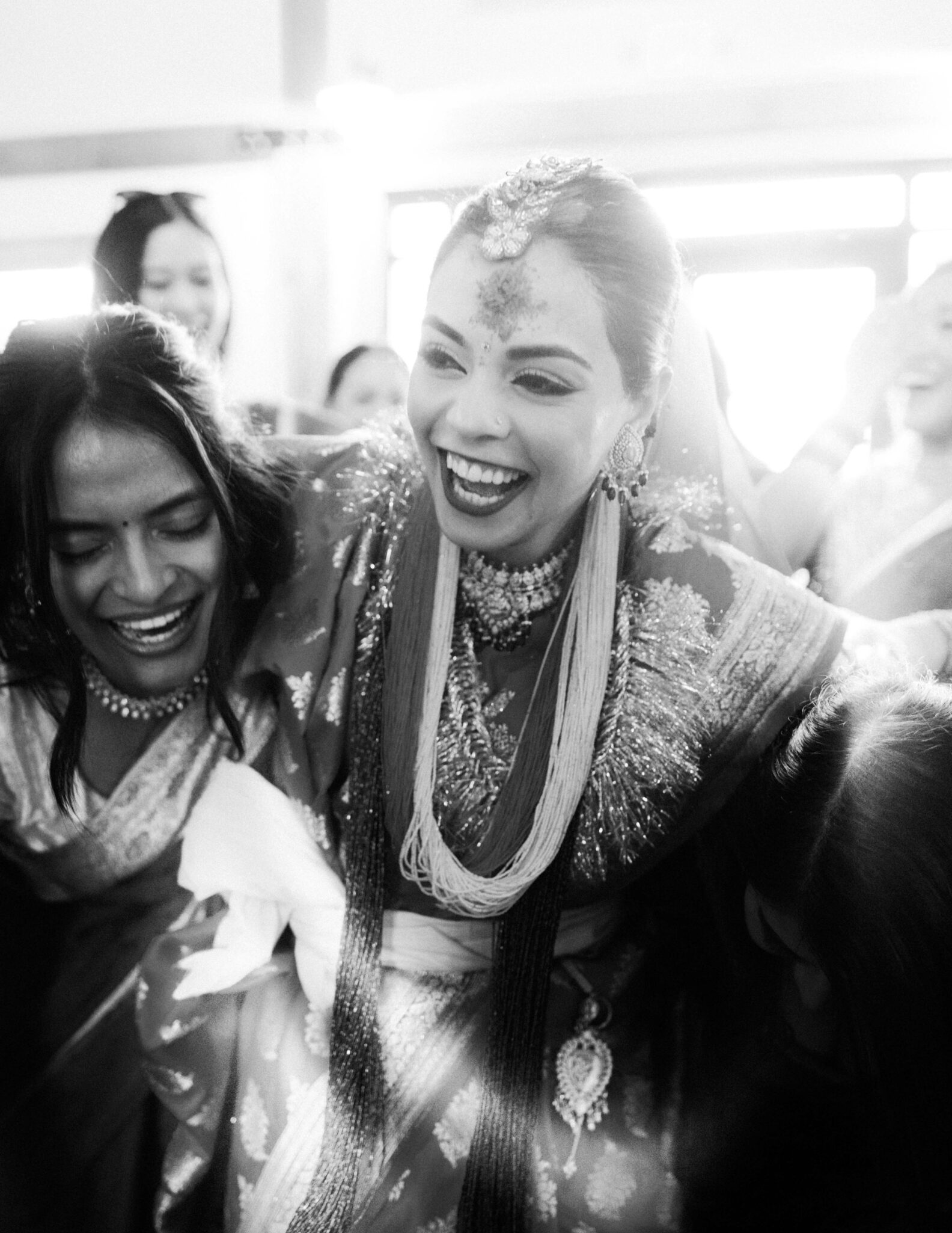 A joyful bride in traditional attire and jewelry smiles and laughs with friends around her. The candid black-and-white photo captures a festive, happy moment.