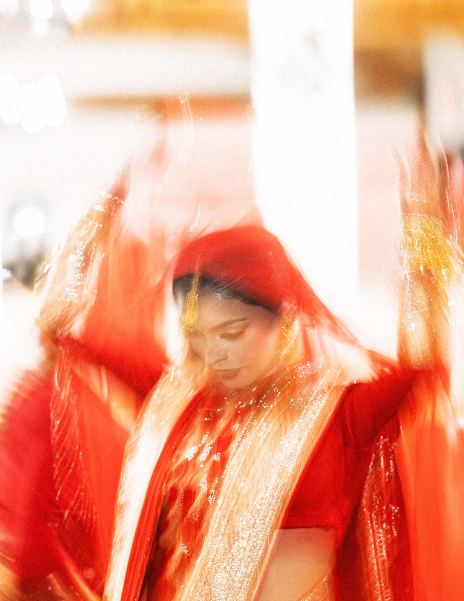 A woman dressed in a vibrant red and gold traditional outfit moves gracefully, her raised arms and flowing attire creating a blurred, dynamic effect that conveys motion and celebration.