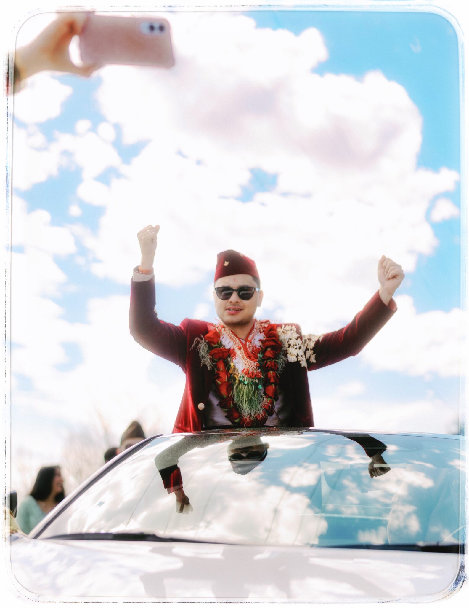 A man wearing sunglasses, maroon suit, and flower garlands stands through a cars sunroof with raised arms, celebrating under a bright, cloudy sky as people watch and someone takes a photo above.