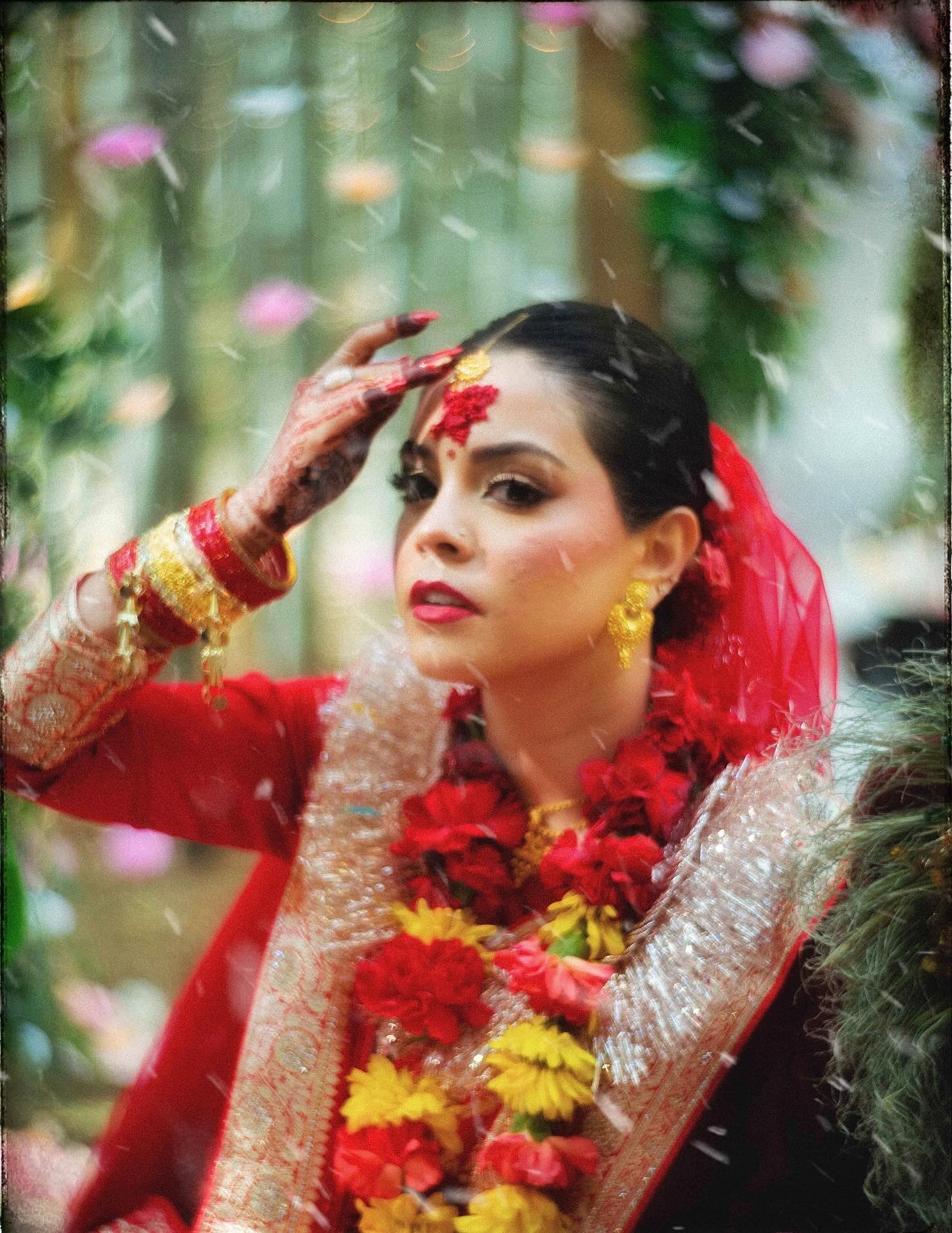 A bride in traditional red attire adorned with gold jewelry and floral garlands touches her forehead, with mehendi on her hand, surrounded by greenery and falling flower petals.
