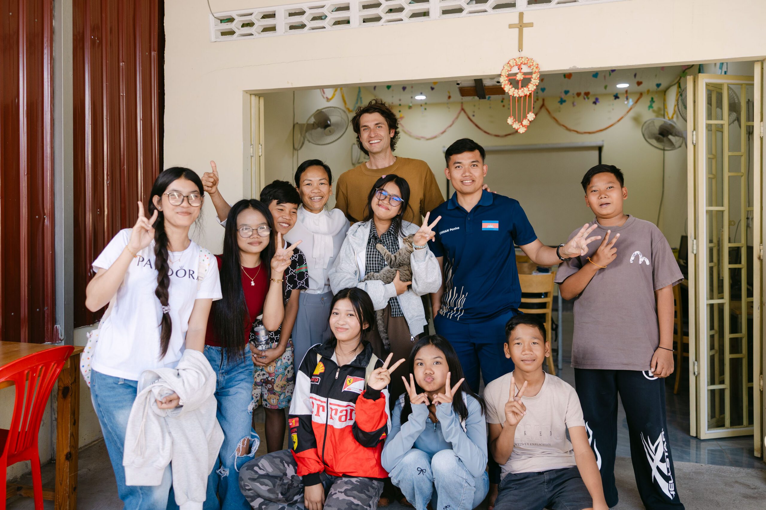A group of people, including adults and children, smile and pose together for a photo inside a decorated room—with a cross and garland behind them—showcasing the spirit of Helping Hand Cambodia as some flash peace signs with their hands.