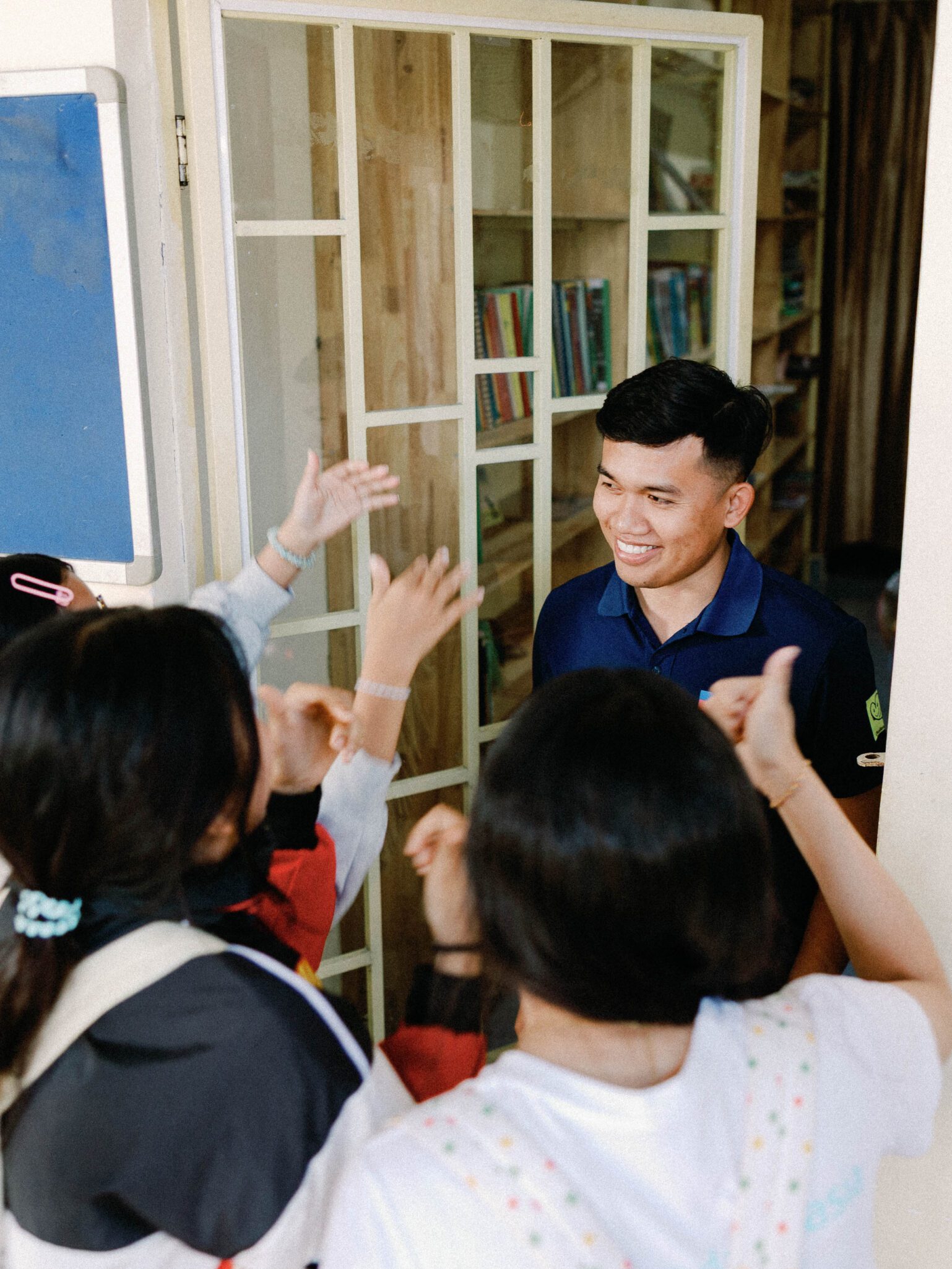 A smiling man stands in a doorway as a group of children excitedly greet him with raised hands inside a room lined with bookshelves.