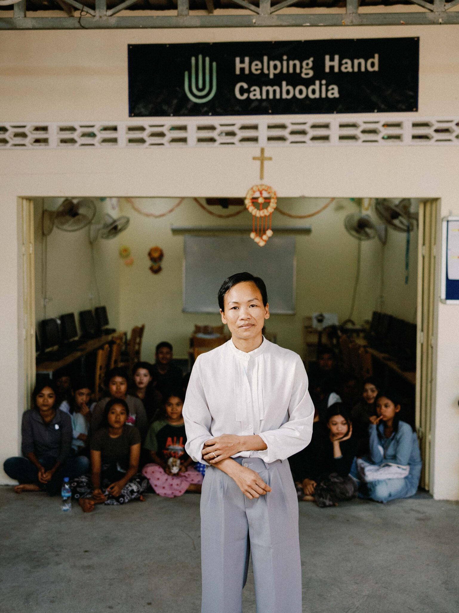 A person in a white shirt stands in front of a group of seated people inside a room. Above the entrance is a sign that reads Helping Hand Cambodia. Several computers line the walls in the background.