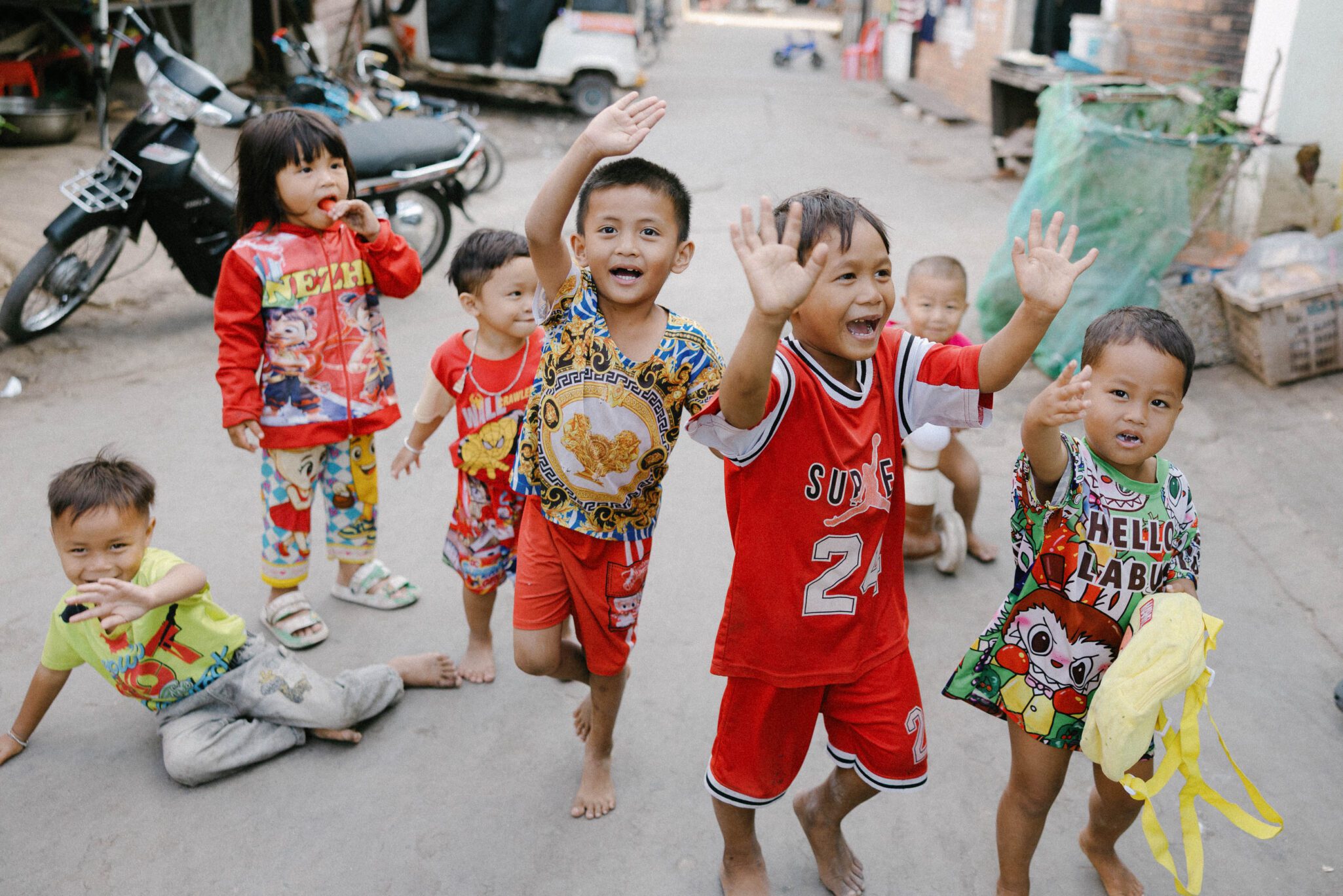 A group of young children outdoors, smiling and waving at the camera. They are wearing colorful clothes and appear happy, standing and kneeling on a paved street with buildings and a parked motorcycle in the background.