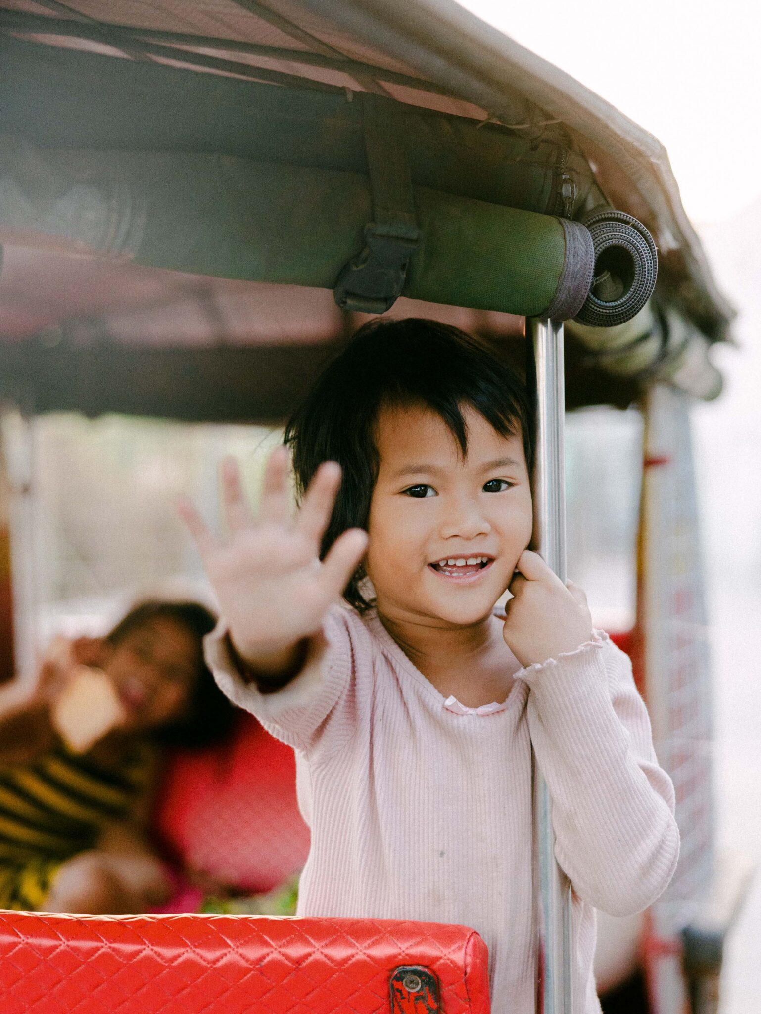 A smiling child in a light pink shirt stands on a red seat, holding a metal pole and waving with one hand. Another child in a striped outfit is sitting in the background, slightly out of focus.