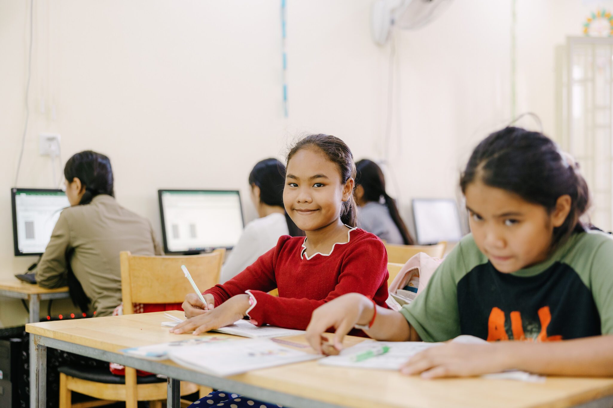 Two girls sit at a classroom desk, writing in notebooks and looking at the camera, while other students work on computers in the background. The classroom is well-lit and organized.