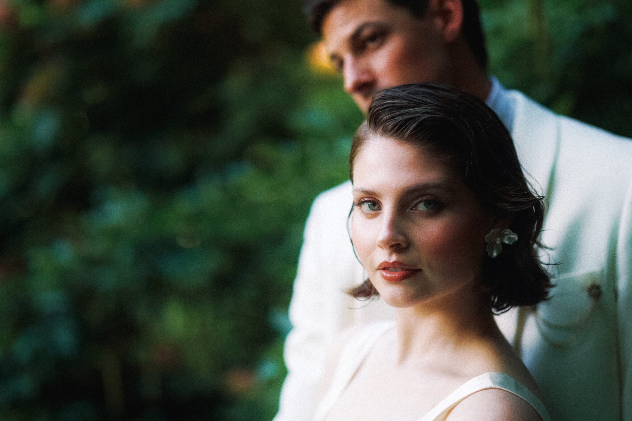 A woman with short dark hair and pearl earrings, wearing a light dress, stands in focus in front of a man in a white suit. The background is blurred greenery.