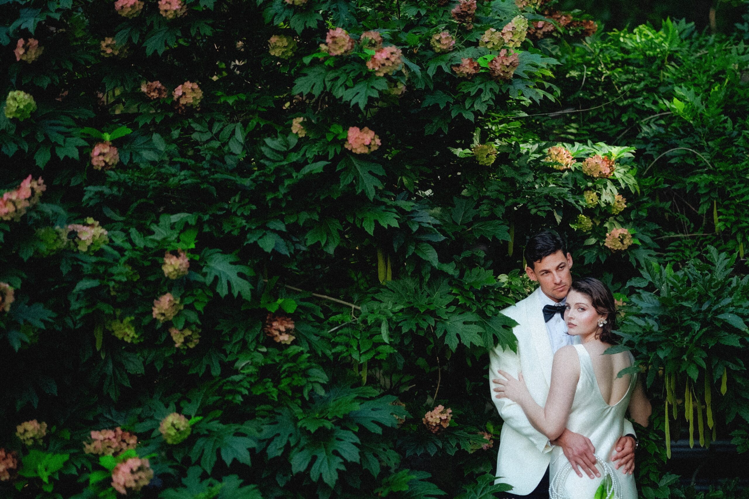 A couple in formal attire stands closely together, surrounded by lush green foliage with pinkish flowers at their elegant Caya Azul Asheville wedding. The man wears a white suit with a black bow tie; the woman wears a white dress and rests her arms on his chest.