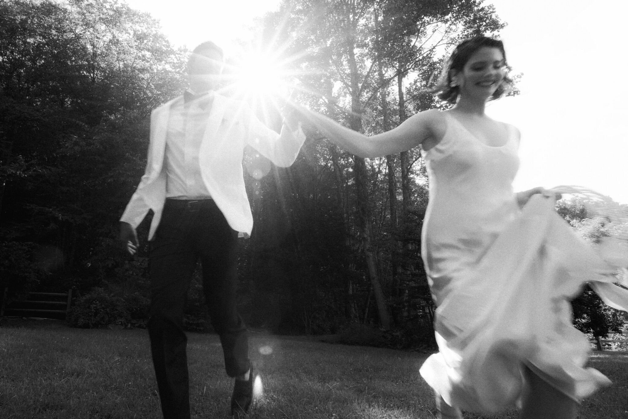 A joyful couple, dressed in formal attire for their Caya Azul Asheville wedding, hold hands and run across a grassy area in sunlight, with trees in the background. The black and white photo captures a bright flare between them.