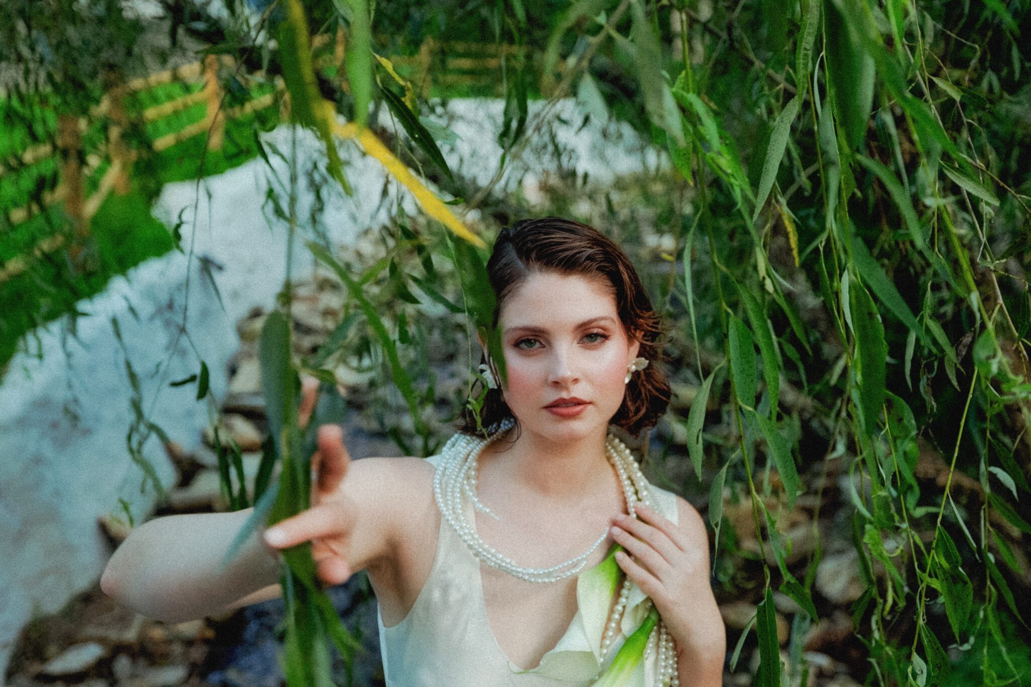 A woman with short brown hair, wearing a white dress and layered pearl necklaces, stands among hanging green willow branches, extending her hand toward the camera with a serene expression. Stones and greenery are visible in the background.
