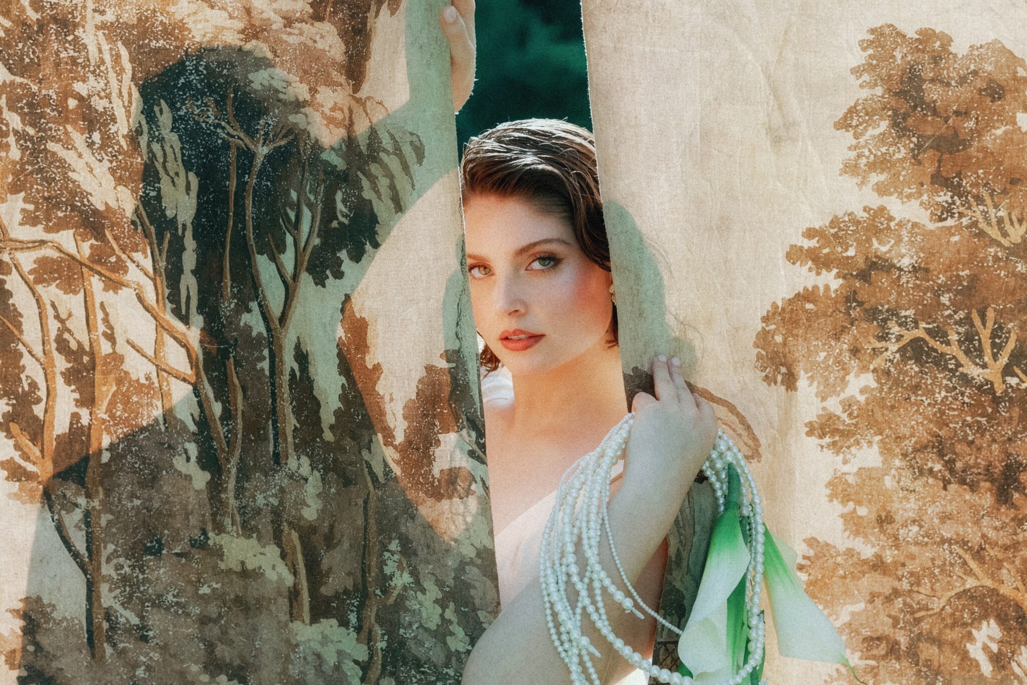 A woman with short brown hair peers through a slit in a vintage, tapestry-like curtain at a Caya Azul Asheville wedding, holding strands of pearls and a green cloth. The curtain’s nature scenes blend with her partially visible face.