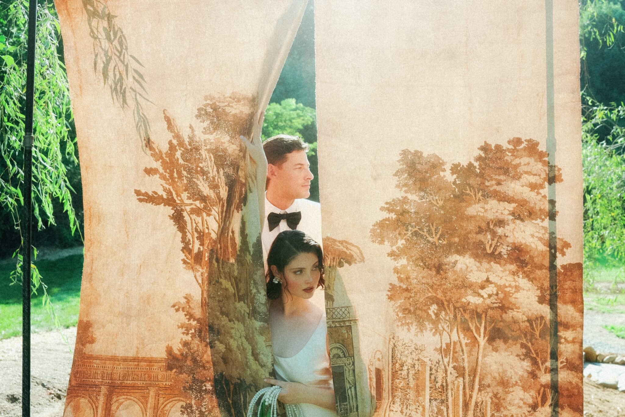 A man in a tuxedo and a woman in a white dress stand behind vintage, nature-themed curtains outdoors, with sunlight filtering through and greenery visible in the background.