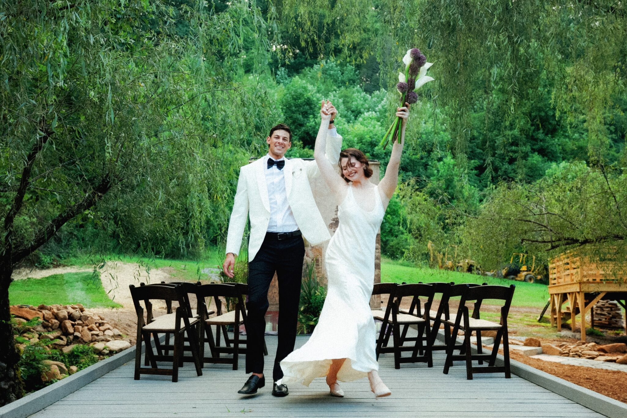 A joyful bride and groom walk hand in hand down an outdoor aisle at their Caya Azul Asheville wedding. The bride raises her bouquet in celebration amid lush greenery, with empty chairs on either side and trees in the background.