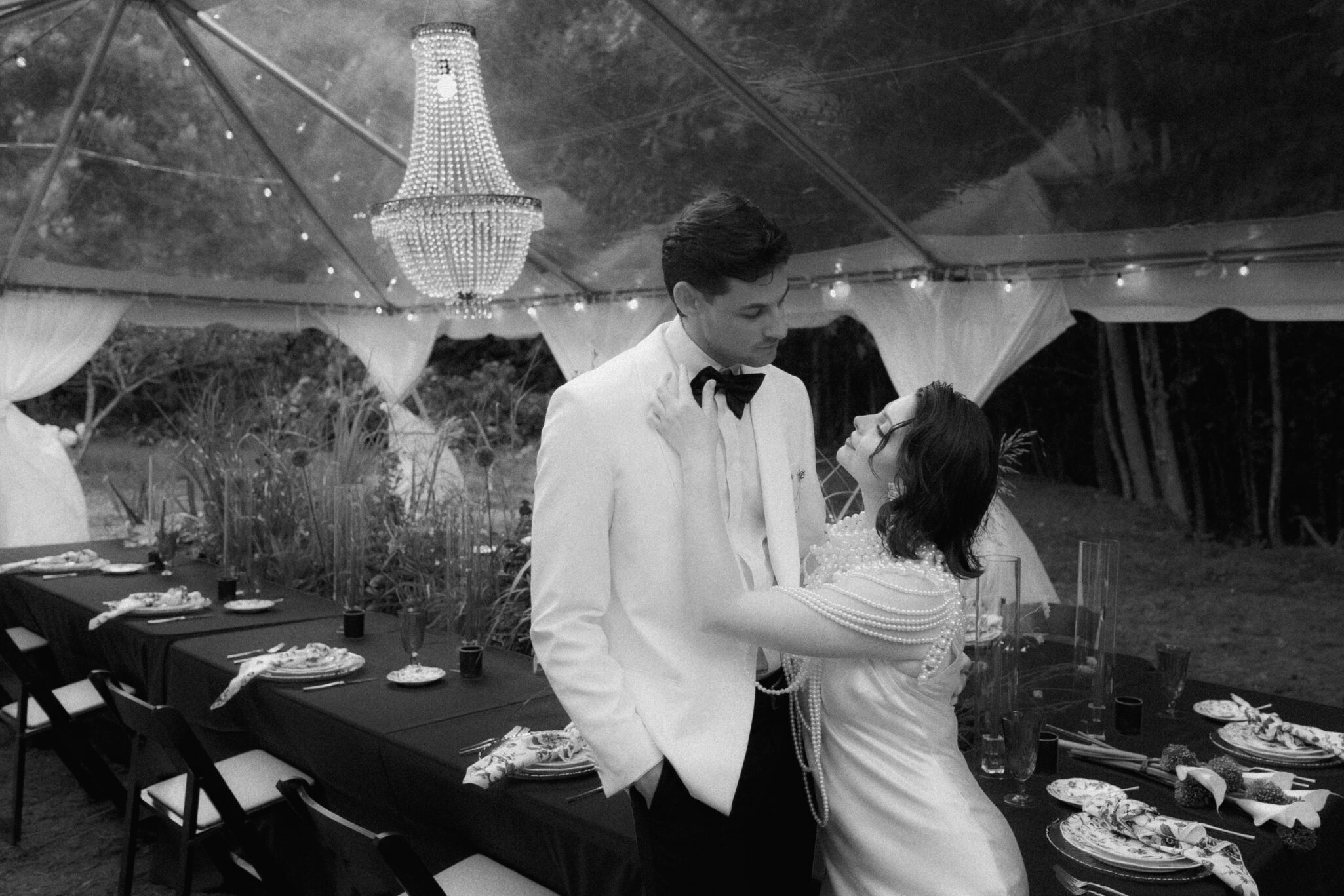 A bride and groom in formal attire stand close together under a clear tent with string lights and a chandelier, gazing at each other beside a long, decorated dining table at an outdoor wedding.