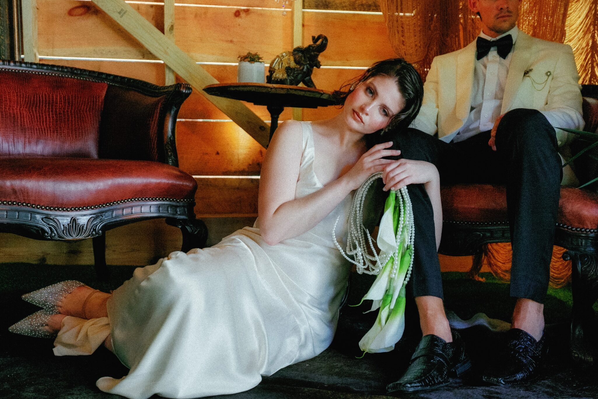 A woman in a white satin dress sits on the floor, leaning against a man's knee at a rustic caya azul Asheville wedding. The man, in a white tuxedo jacket and black pants, relaxes on a vintage red sofa amid wooden walls and warm lighting.
